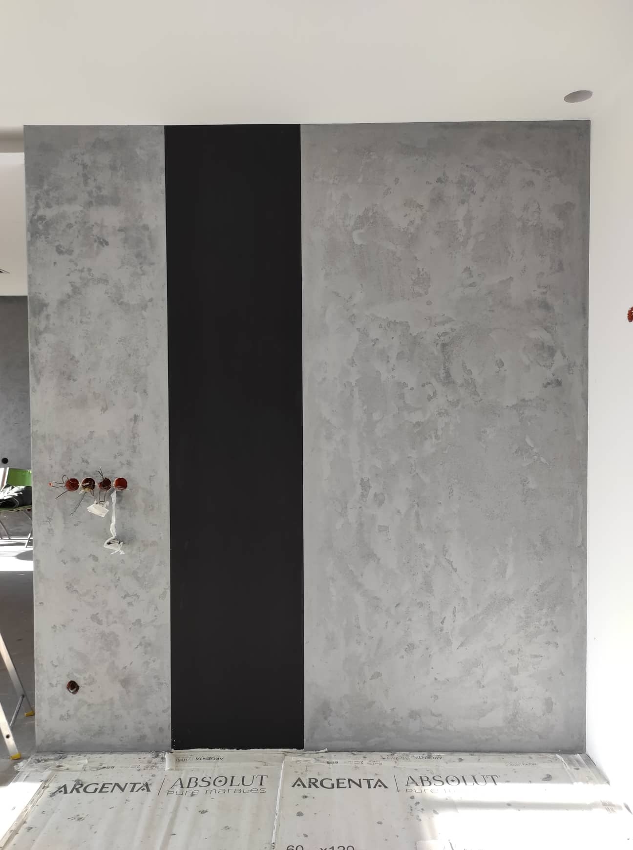 Ściana z dekoracyjną szarą strukturą tynku i pionowym pasem czarnej farby, widoczne wystające kable elektryczne i zabezpieczona podłoga z kartonami.