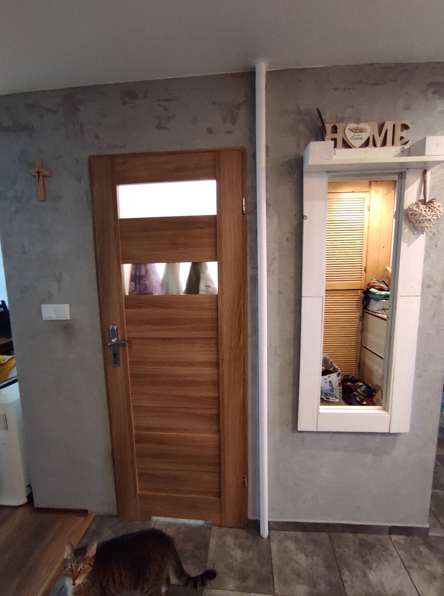 Ściana o szarej, surowej fakturze z drewnianymi drzwiami z przeszkleniem, białym lustrem z półką i dekoracjami, uzupełniona o krzyż na ścianie i kota w dolnej części kadru.