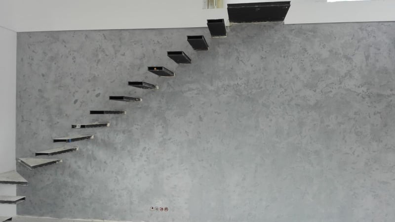 Ściana z dekoracyjnym tynkiem w odcieniach szarości, obok schody wspornikowe z czarnymi elementami mocującymi do ściany.