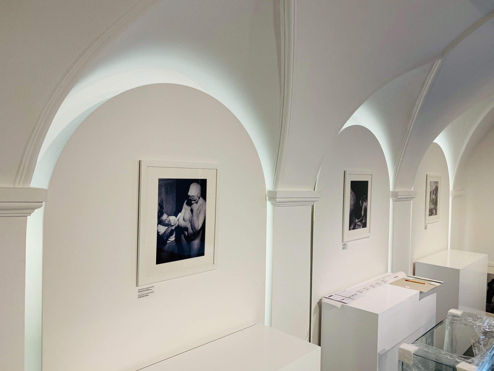 Wnętrze galerii sztuki z białymi ścianami, łukowym sufitem i czarno-białymi fotografiami w ramkach, umieszczonymi na ścianach. Na pierwszym planie białe postumenty i stoliki.