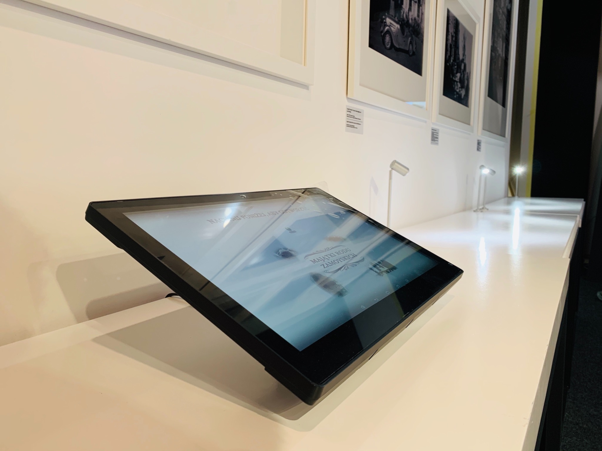 Interaktywny ekran dotykowy na białym postumencie w muzeum lub galerii, prezentujący informacje o Majątkach Rodu Zamoyskich, z czarno-białymi fotografiami w tle.