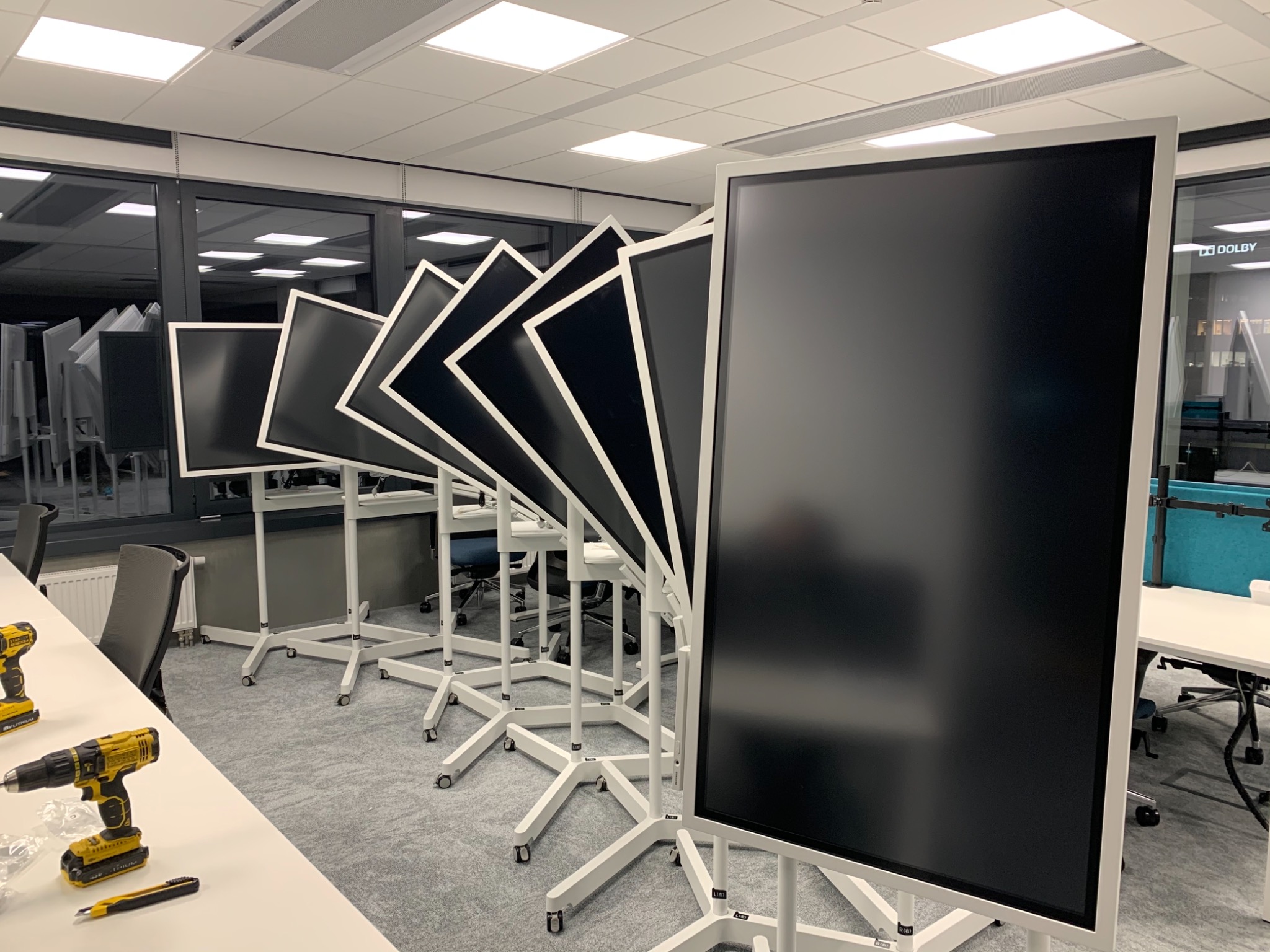 Rząd ustawionych w rzędzie, jeszcze niepodłączonych, dużych, czarnych ekranów na białych, mobilnych stojakach w jasnym biurze, obok wiertarki i nożyk na stole, z widocznymi oknami i biurami w tle.