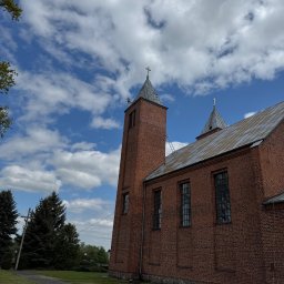 DACH- BRUK ZDZISŁAW KOŁTUNIUK - Ceglany kościół z metalowym dachem pod błękitnym niebem. Drabina oparta o wieżę. Widok z dołu, zieleń w tle. Prace na wysokości.