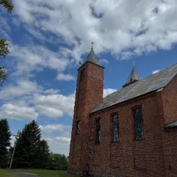 Ceglany kościół z metalowym dachem pod błękitnym niebem. Drabina oparta o wieżę. Widok z dołu, zieleń w tle. Prace na wysokości.