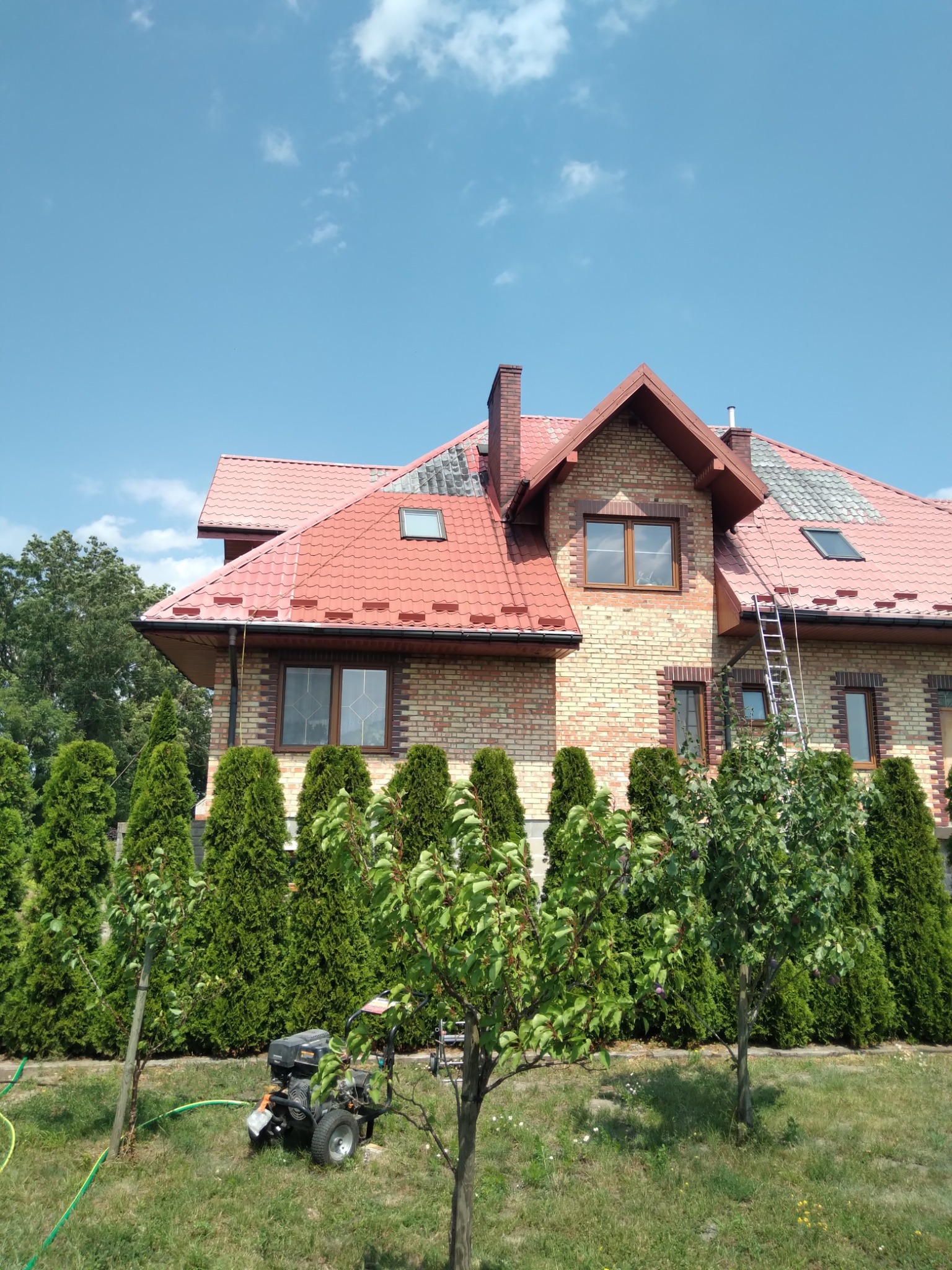 Dwupiętrowy dom z czerwonym dachem z cegły, częściowo pomalowanym na szaro, z drabiną opartą o ścianę i myjką ciśnieniową na trawniku przed budynkiem.
