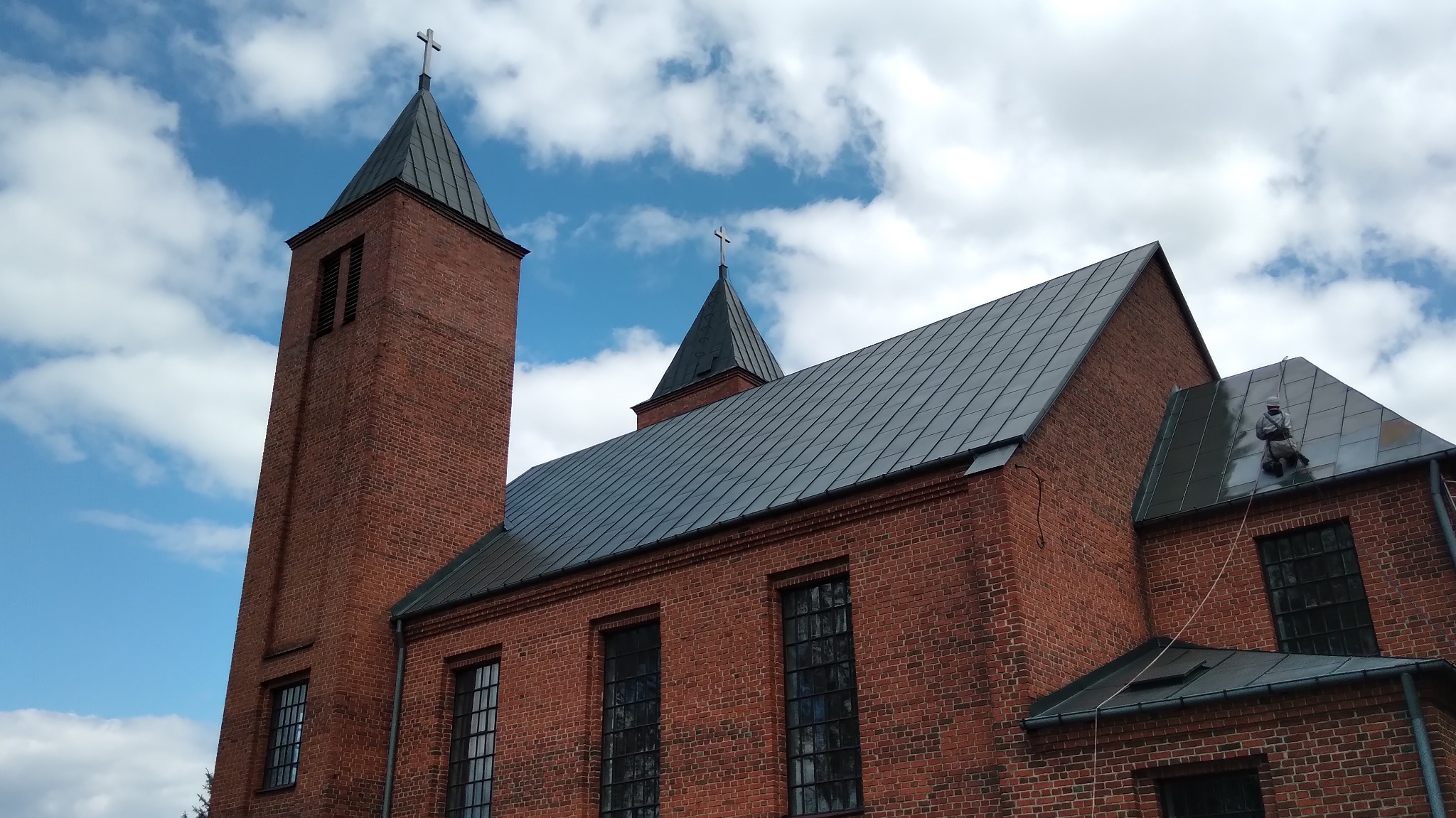 Ceglany kościół z ciemnym dachem pod błękitnym niebem. Na dachu, zabezpieczony liną, pracuje malarz w kombinezonie, odświeżając jego kolor.