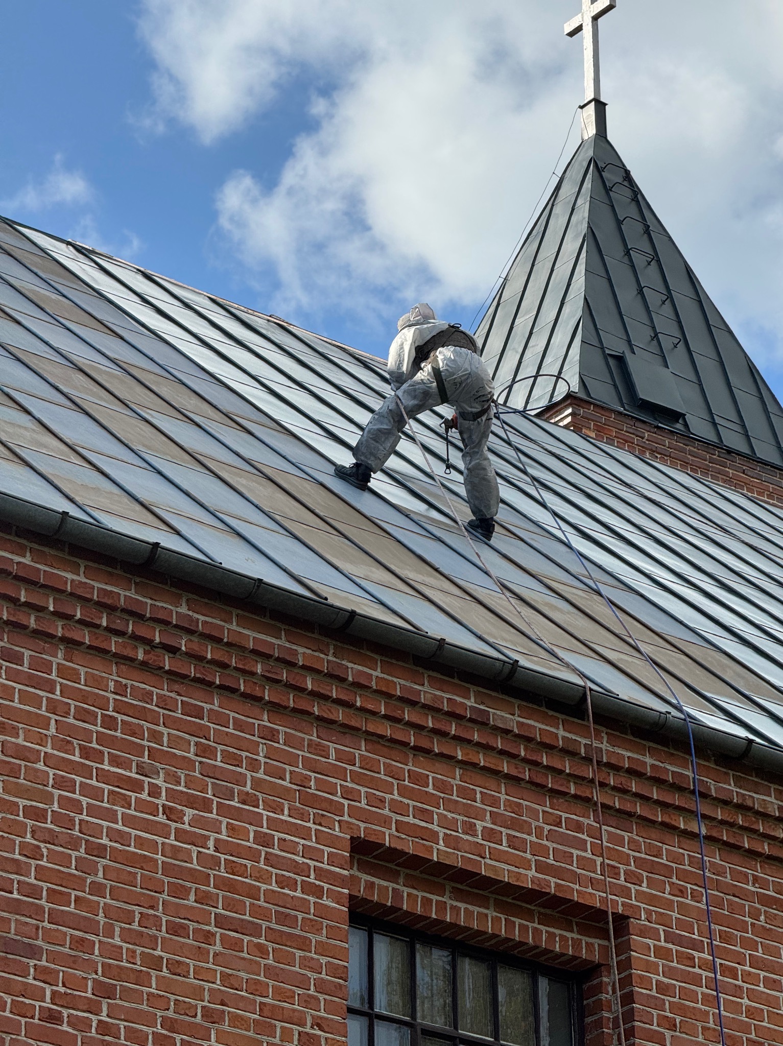 Pracownik w kombinezonie na metalowym dachu kościoła, zabezpieczony linami, z krzyżem na szczycie. Ceglana ściana budynku w tle.