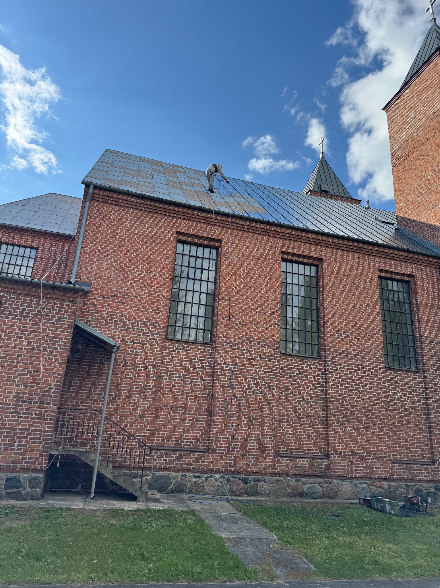 Pracownik na dachu kościoła z cegły, wykonujący prace renowacyjne. Widoczne narzędzia i puszki z farbą. Błękitne niebo z chmurami w tle.