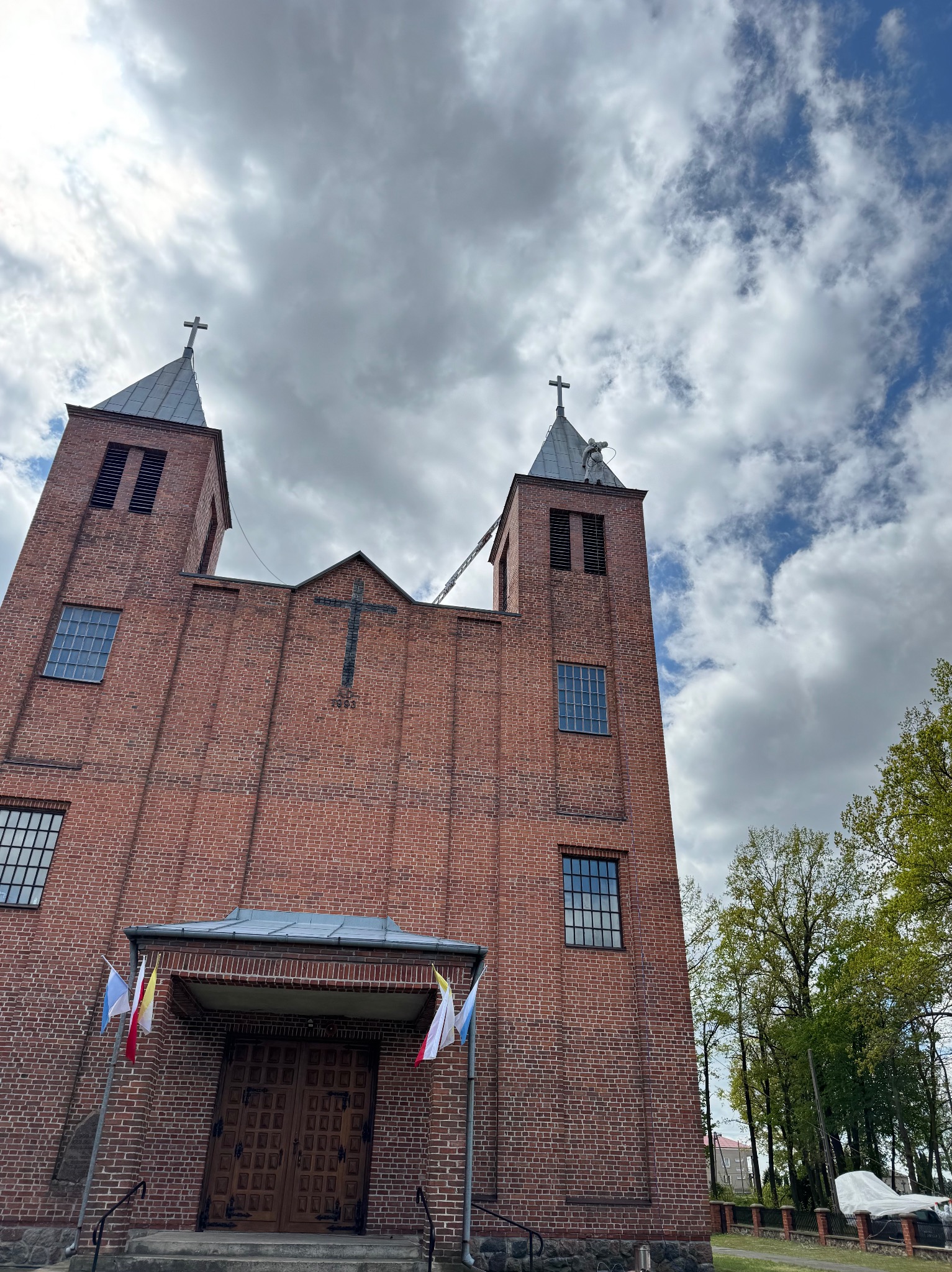 Ceglany kościół z dwiema wieżami i krzyżem na fasadzie, z flagami przed wejściem, na tle pochmurnego nieba. Widok z dołu.