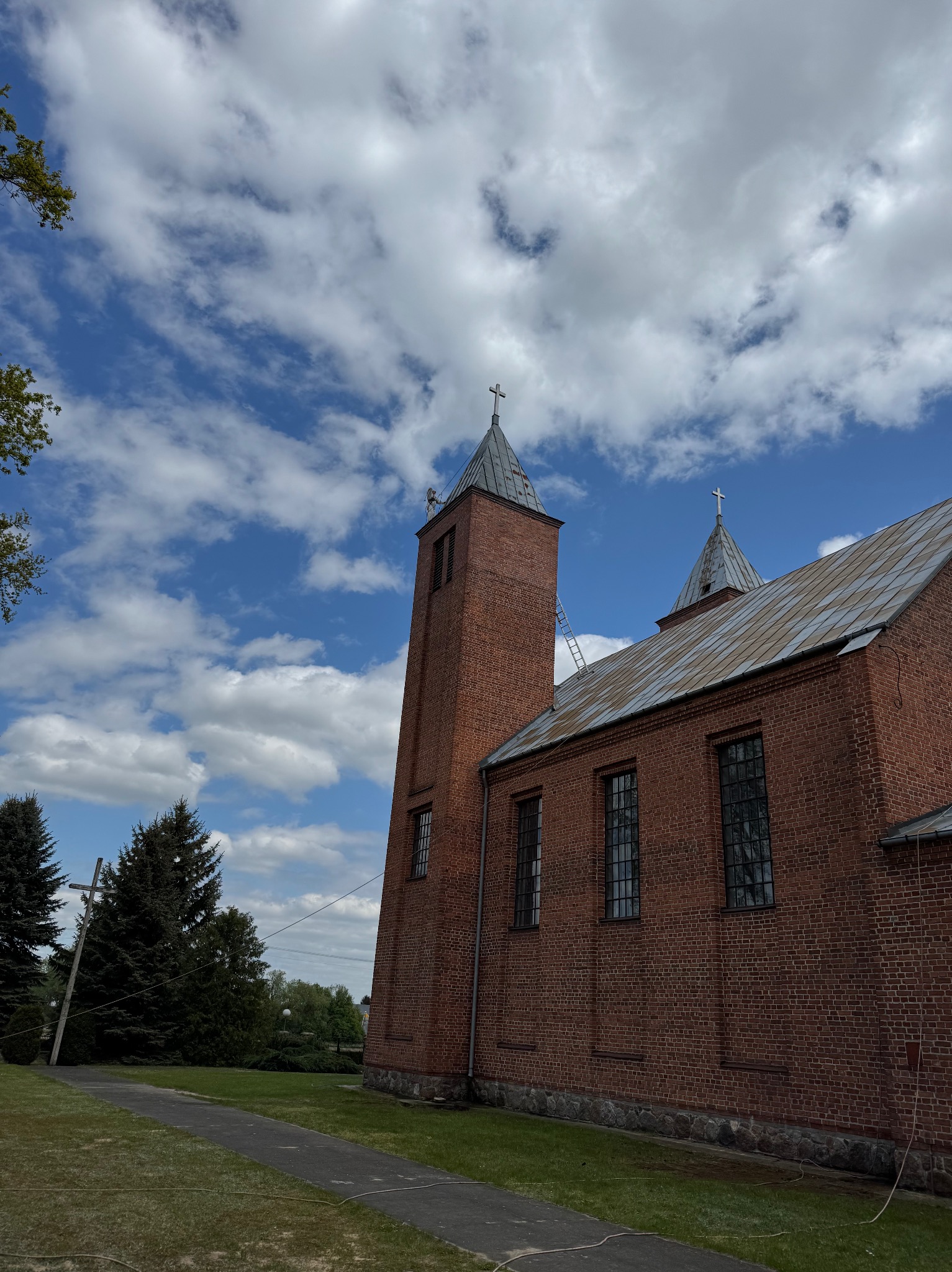 Ceglany kościół z metalowym dachem pod błękitnym niebem. Drabina oparta o wieżę. Widok z dołu, zieleń w tle. Prace na wysokości.