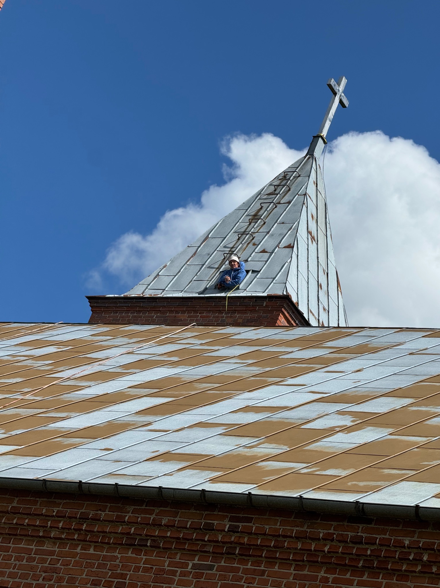 Pracownik w kasku na dachu kościoła z blachy, częściowo pomalowanym na brązowo. W tle niebieskie niebo i krzyż na szczycie wieżyczki.