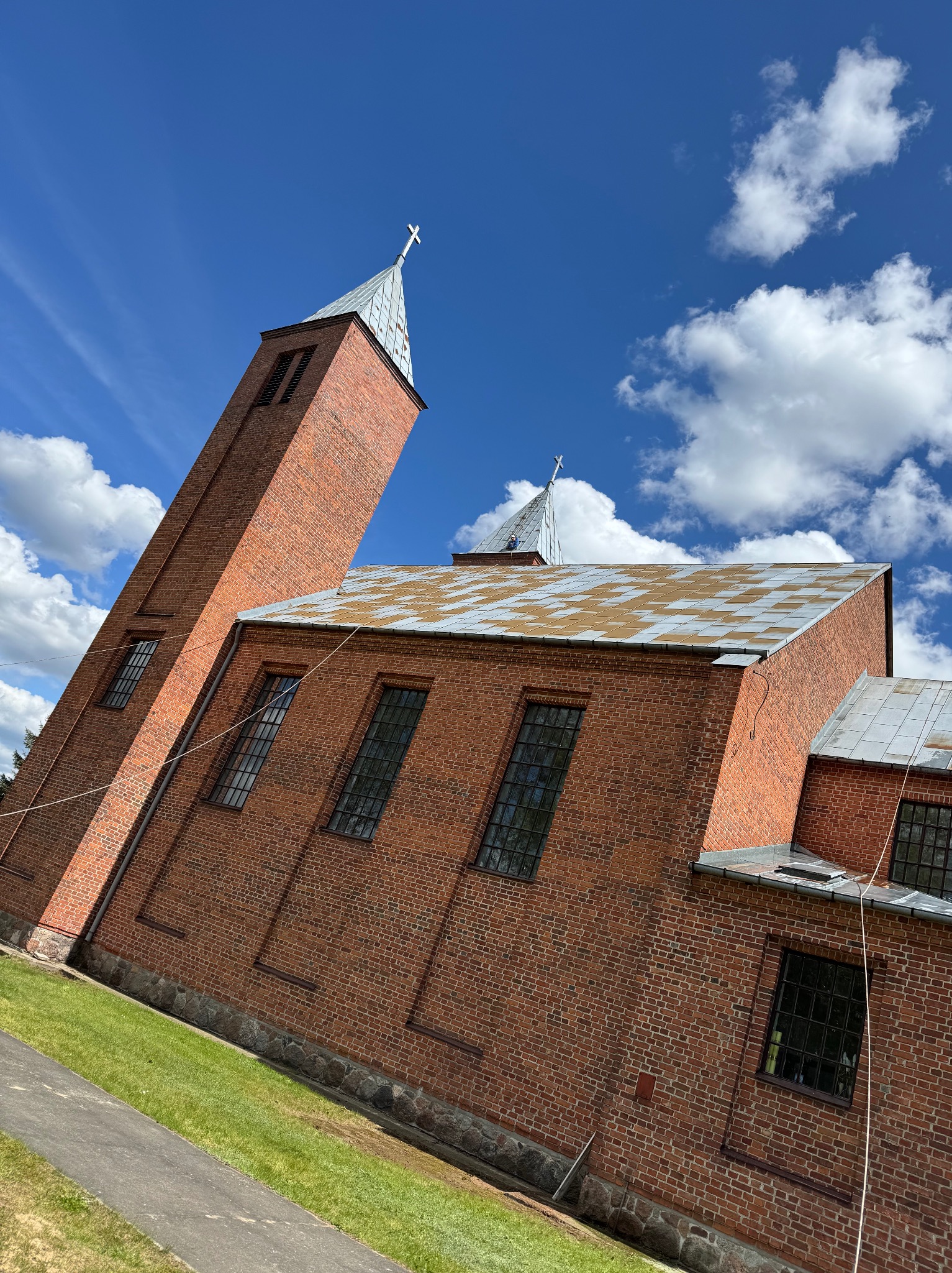 Ceglany kościół z wieżą, widziany pod kątem, z fragmentem dachu w szachownicę i błękitnym niebem w tle. Widoczne okna i trawnik.