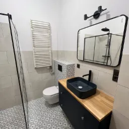 Nowoczesna łazienka z czarną umywalką nablatową, szafką z drewnianym blatem, lustrem w czarnej ramie i kabiną prysznicową ze szklaną ścianką.