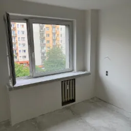 Pokój po nałożeniu gładzi, przygotowany do dalszych prac wykończeniowych, widoczne zabezpieczone okno i grzejnik, surowe gniazdka elektryczne.