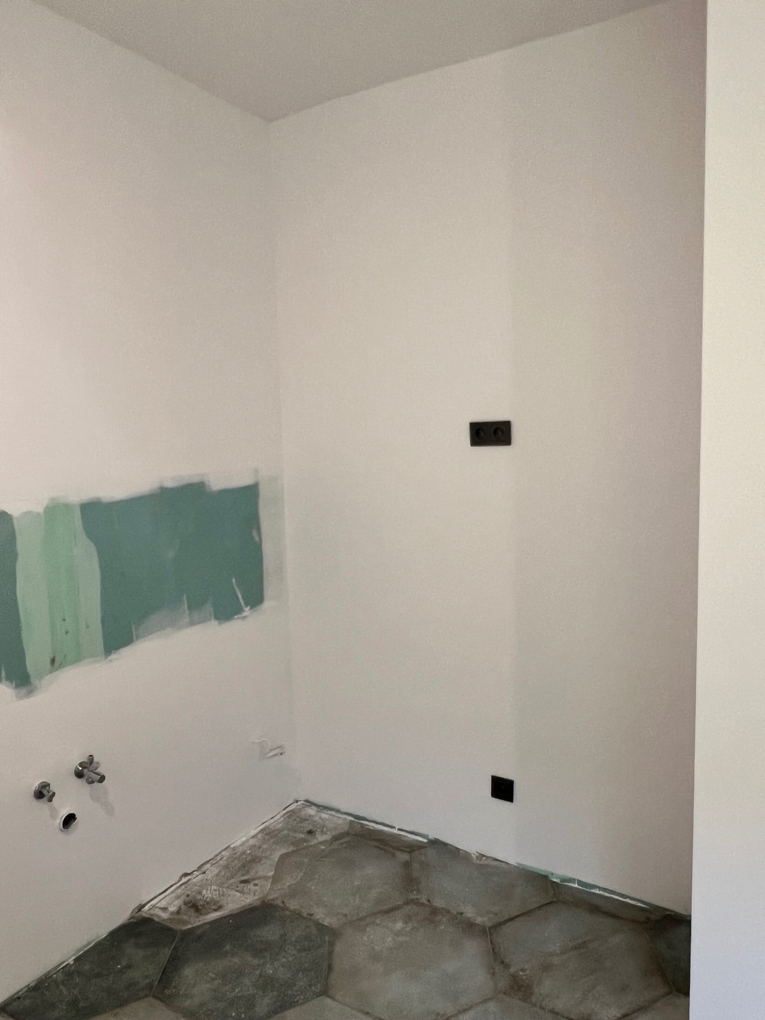Przygotowanie ścian do prac wykończeniowych: fragment ściany z nałożoną warstwą szpachli w odcieniach zieleni, widoczne instalacje hydrauliczne i elektryczne, oraz fragment podłogi wyłożonej...