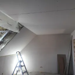 Poddasze w trakcie prac wykończeniowych: ocieplenie wełną mineralną widoczne za płytami gipsowo-kartonowymi, metalowa drabina na posadzce z resztkami materiałów.