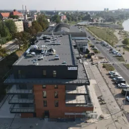 Widok z góry na nowo wybudowany budynek mieszkalny z balkonami, przylegający do chodnika z kostki brukowej i ulicy z zaparkowanymi samochodami, w tle panorama miasta i rzeka.