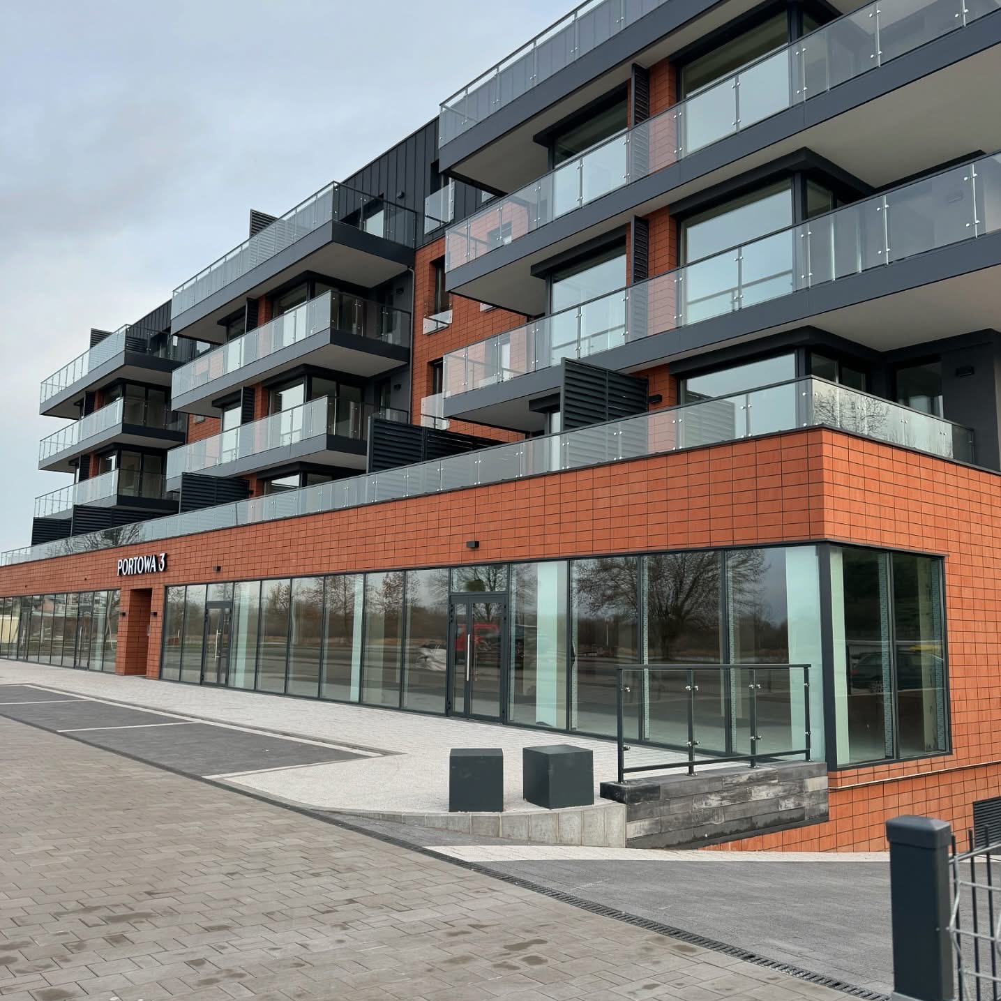 Nowoczesny budynek apartamentowo-usługowy z balkonami z szklanymi balustradami, elewacją z cegły klinkierowej i chodnikiem z kostki brukowej.