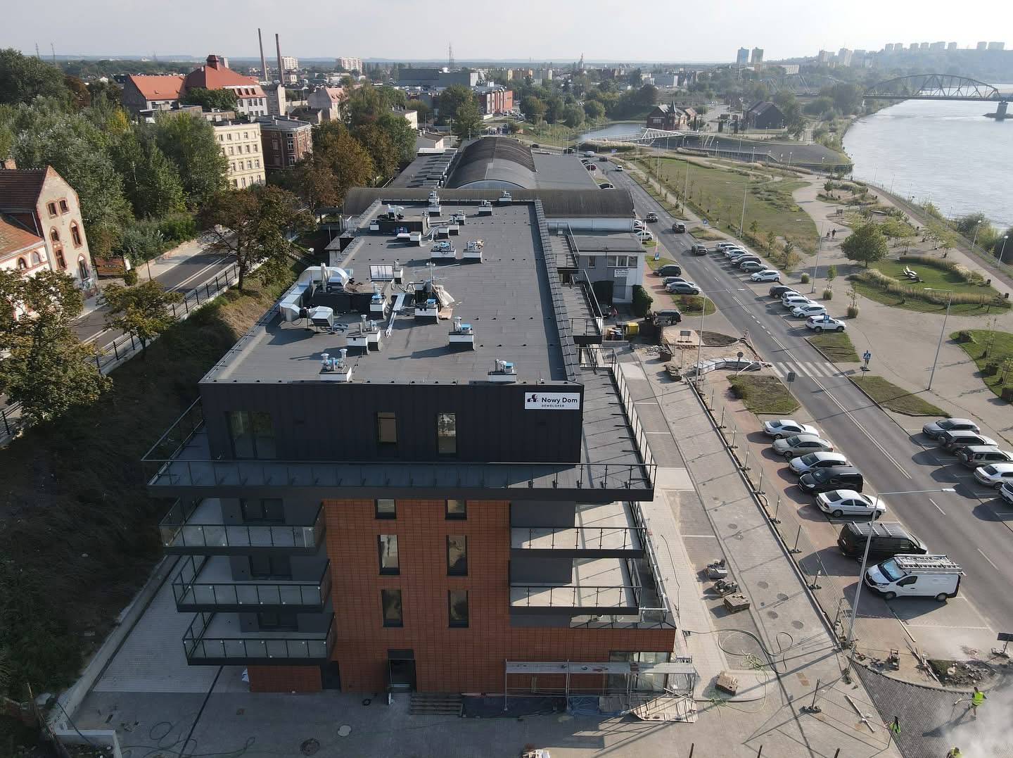 Widok z góry na nowo wybudowany budynek mieszkalny z balkonami, przylegający do chodnika z kostki brukowej i ulicy z zaparkowanymi samochodami, w tle panorama miasta i rzeka.