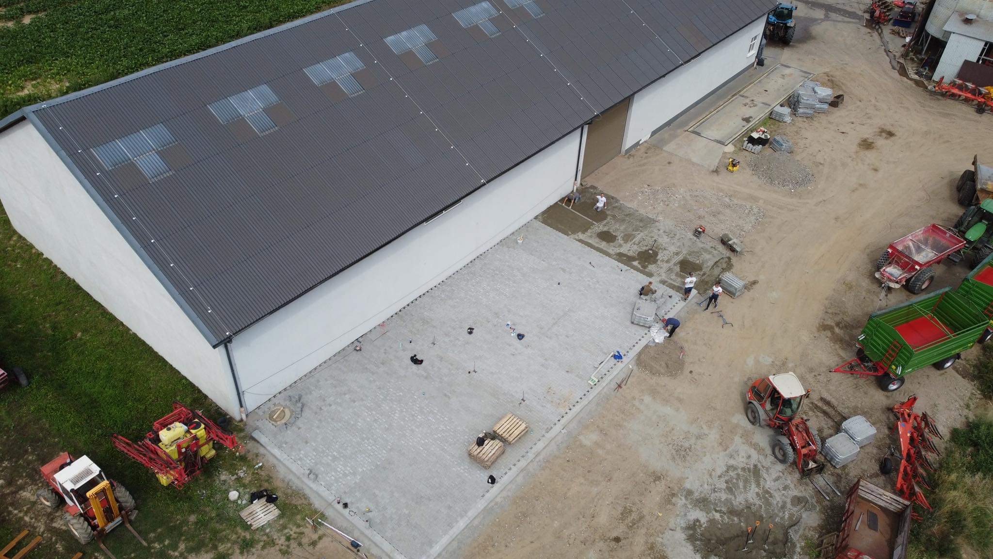 Widok z drona na plac budowy z ułożoną częścią szarej kostki brukowej przed budynkiem magazynowym; pracownicy w jasnych ubraniach układają kolejne elementy, a w tle widoczne maszyny rolnicze i pole...