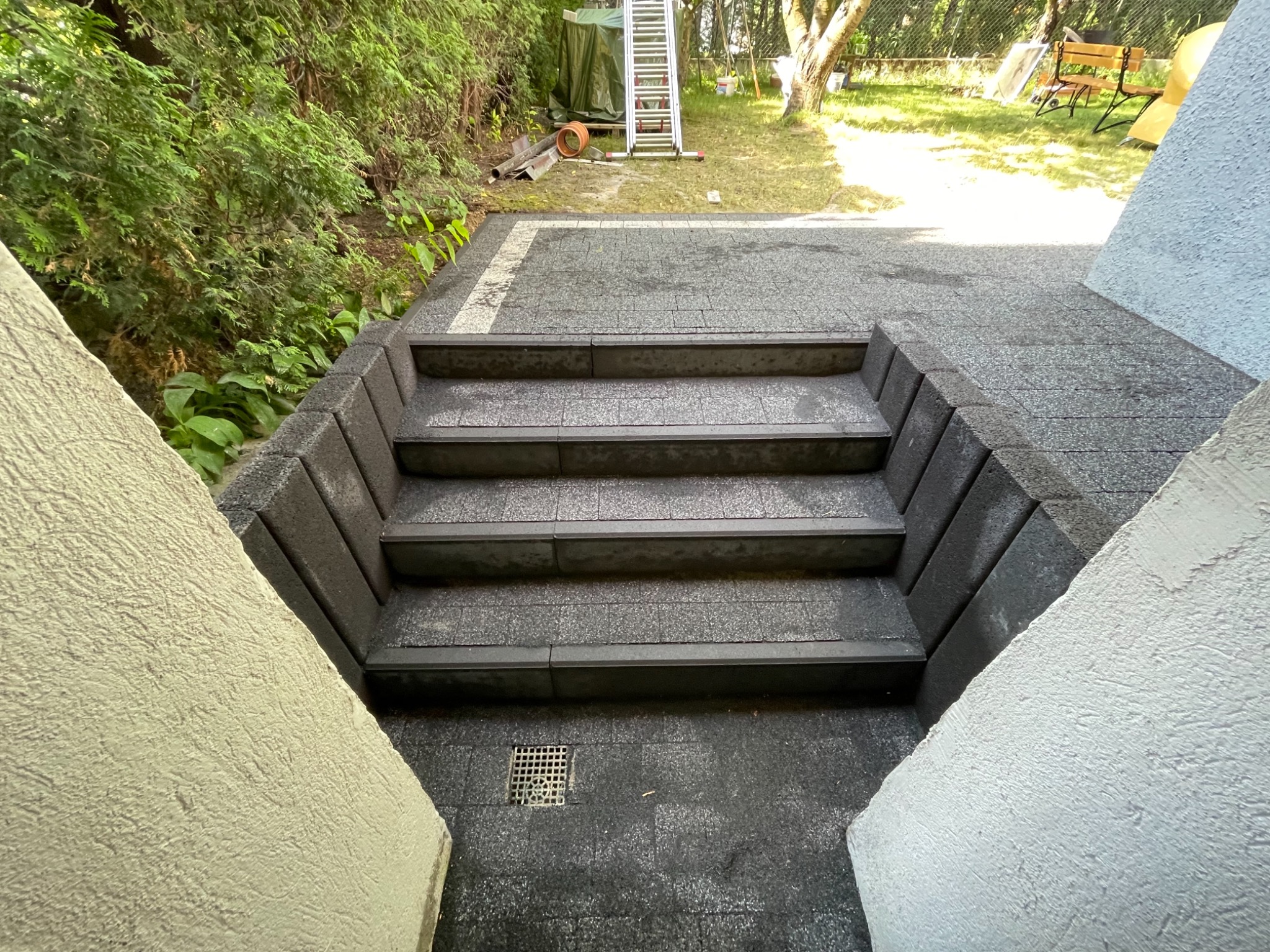 Schody z kostki brukowej prowadzące na taras, z widoczną kratką odpływową i fragmentem ogrodu w tle.
