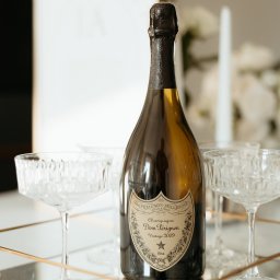 Paweł Ludwikowski - Fotografia - Butelka szampana Dom Pérignon Vintage 2009 na złotej tacy z kryształowymi kieliszkami, białe kwiaty w tle. Elegancka kompozycja, idealna na uroczystość.