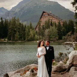 Para młoda w eleganckich strojach pozuje nad jeziorem Morskie Oko, z widokiem na góry i budynek w tle. Romantyczna sceneria, naturalne światło, profesjonalna fotografia.