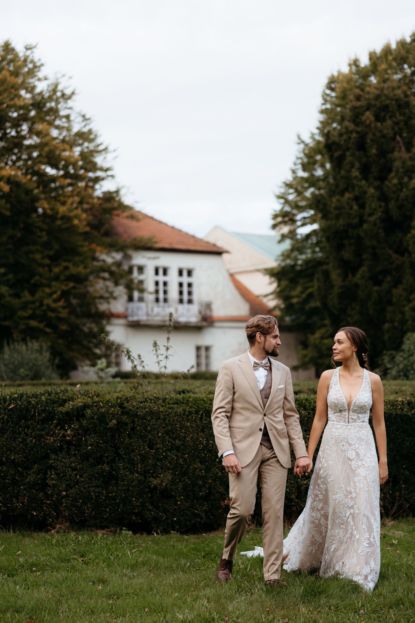 Elegancka para młoda, on w beżowym garniturze z muchą, ona w koronkowej sukni, spacerują po zielonym trawniku na tle klasycznego budynku.