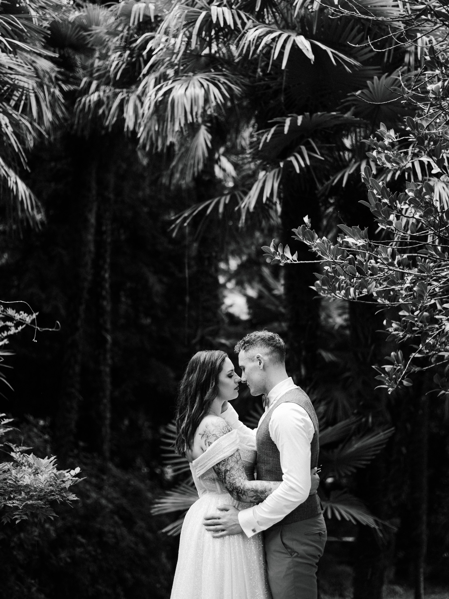 Czarno-białe ujęcie pary młodej w objęciach, z tatuażami na ramionach panny młodej, na tle egzotycznej roślinności podczas sesji plenerowej.