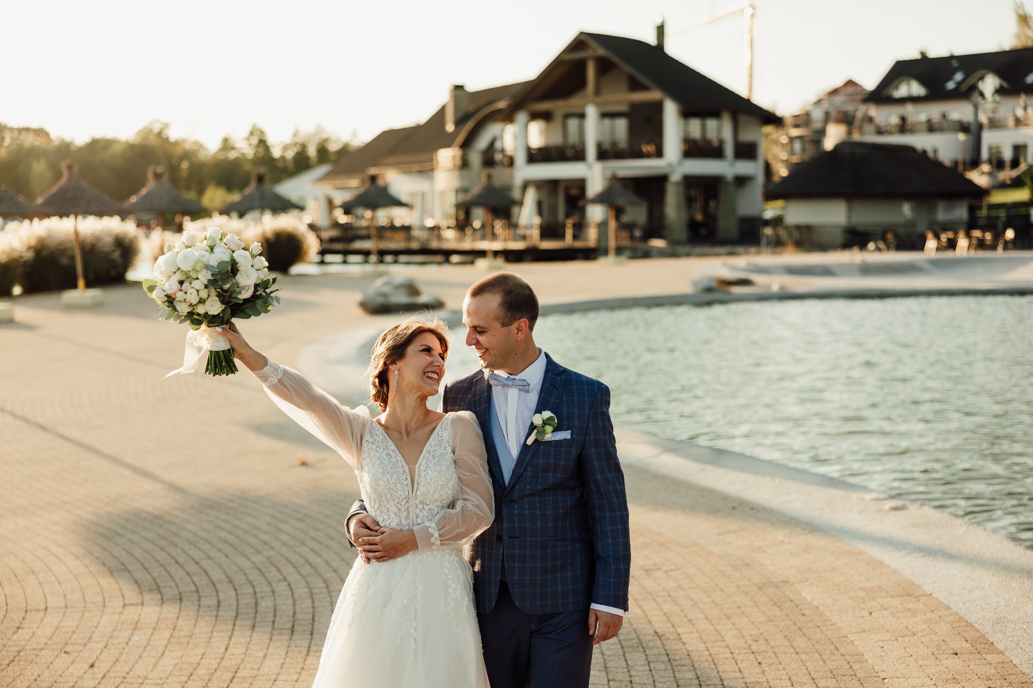 Uśmiechnięta para młoda w plenerze, ona z bukietem, on w granatowym garniturze w kratę, jezioro i budynek w tle, ciepłe światło.