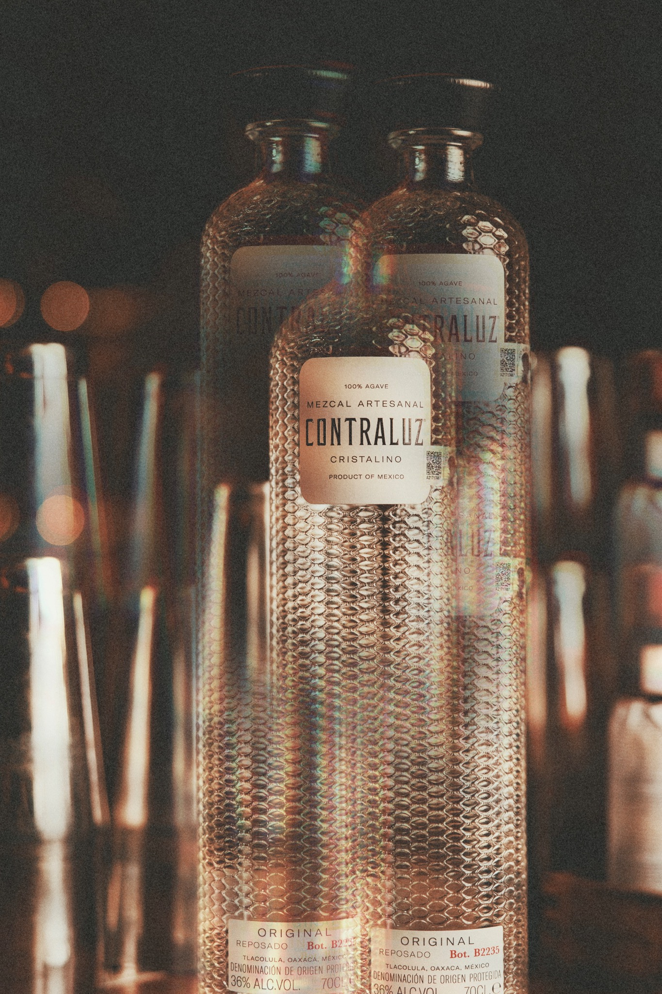 Dwie butelki Contraluz Cristalino Mezcal Reposado z fakturą przypominającą skórę węża, ustawione na tle baru z rozmazanymi kieliszkami i shakerami.