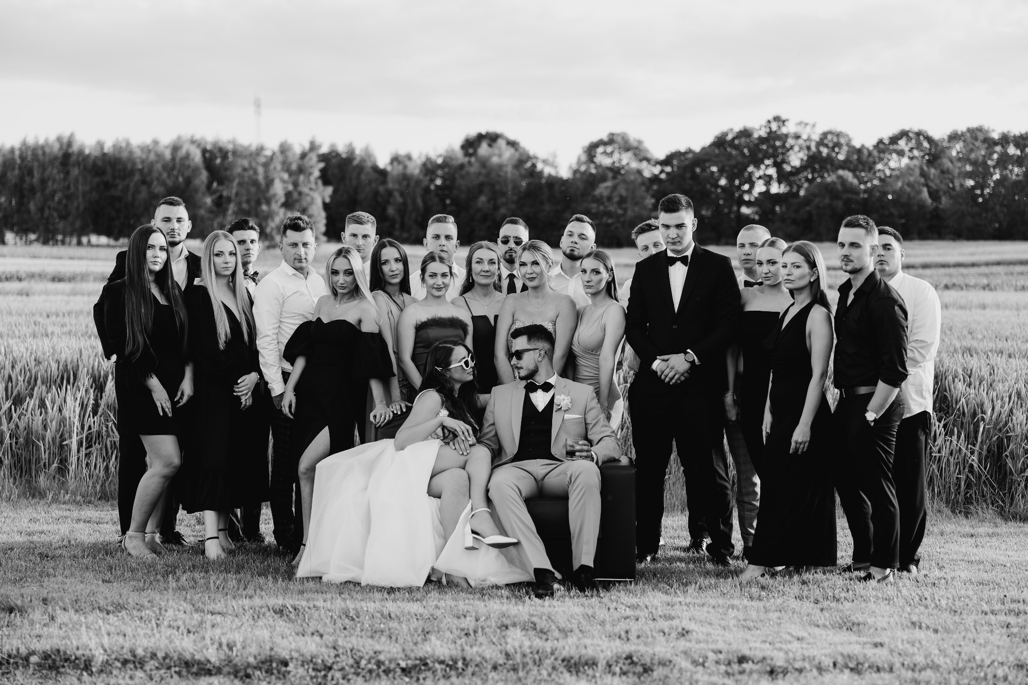 Czarno-białe zdjęcie ślubne: Para młoda siedzi na kanapie w polu, otoczona przez gości weselnych. Nietypowe ujęcie, eleganckie stroje, lekko pochmurne niebo.