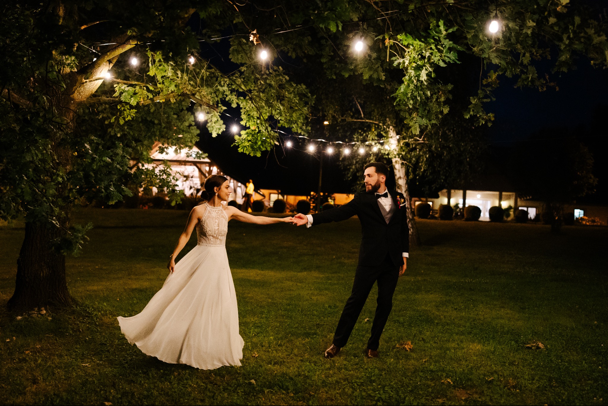 Para młoda trzymająca się za ręce pod drzewem z girlandami świetlnymi w nocy. Bride w sukni ślubnej z koronką, pan młody w czarnym smokingu i muszce.