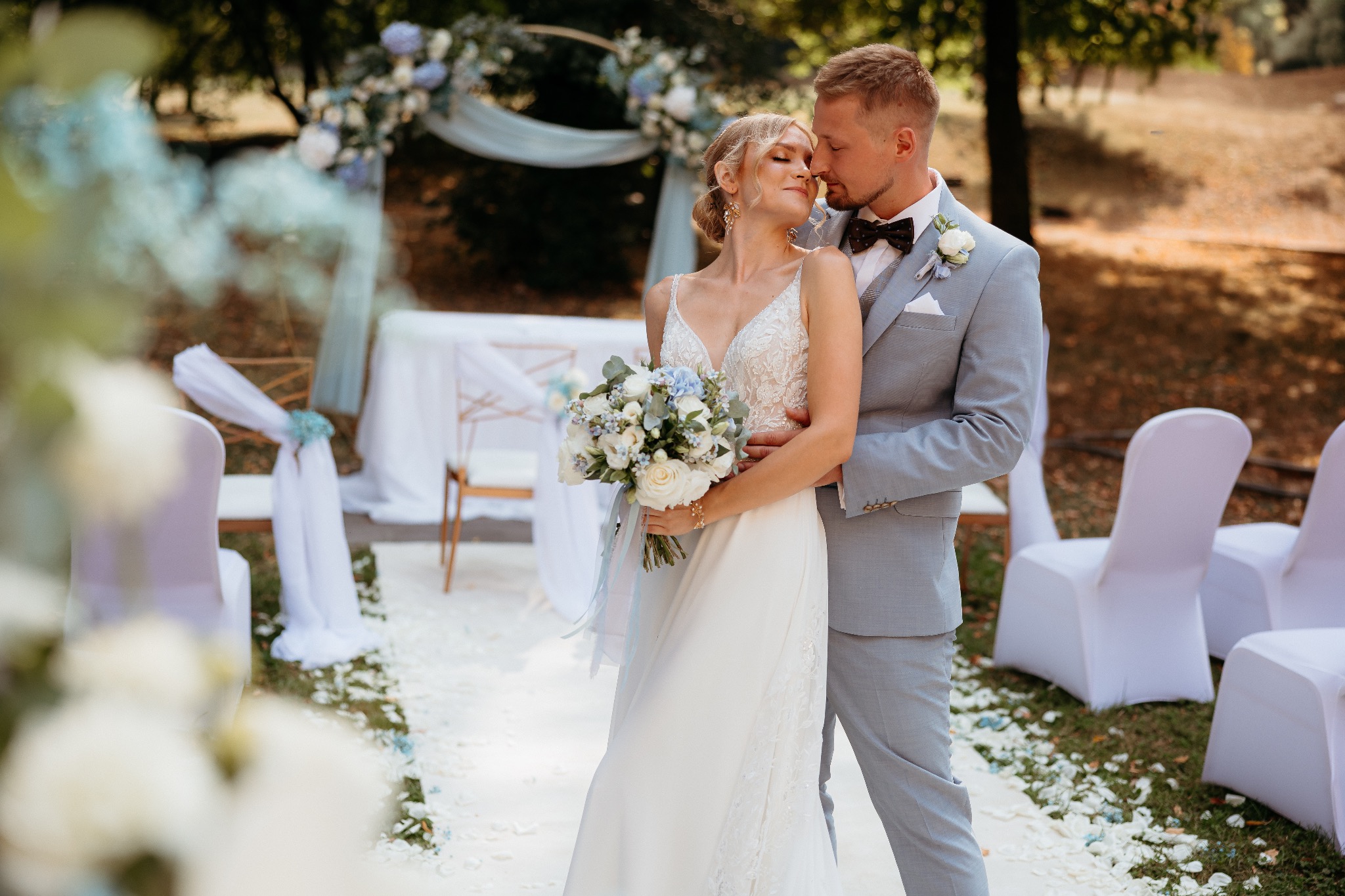 Para młoda w objęciach na tle udekorowanej ceremonii ślubnej. Panna młoda trzyma bukiet z białych róż i niebieskich akcentów, pan młody w szarym garniturze z muszką.