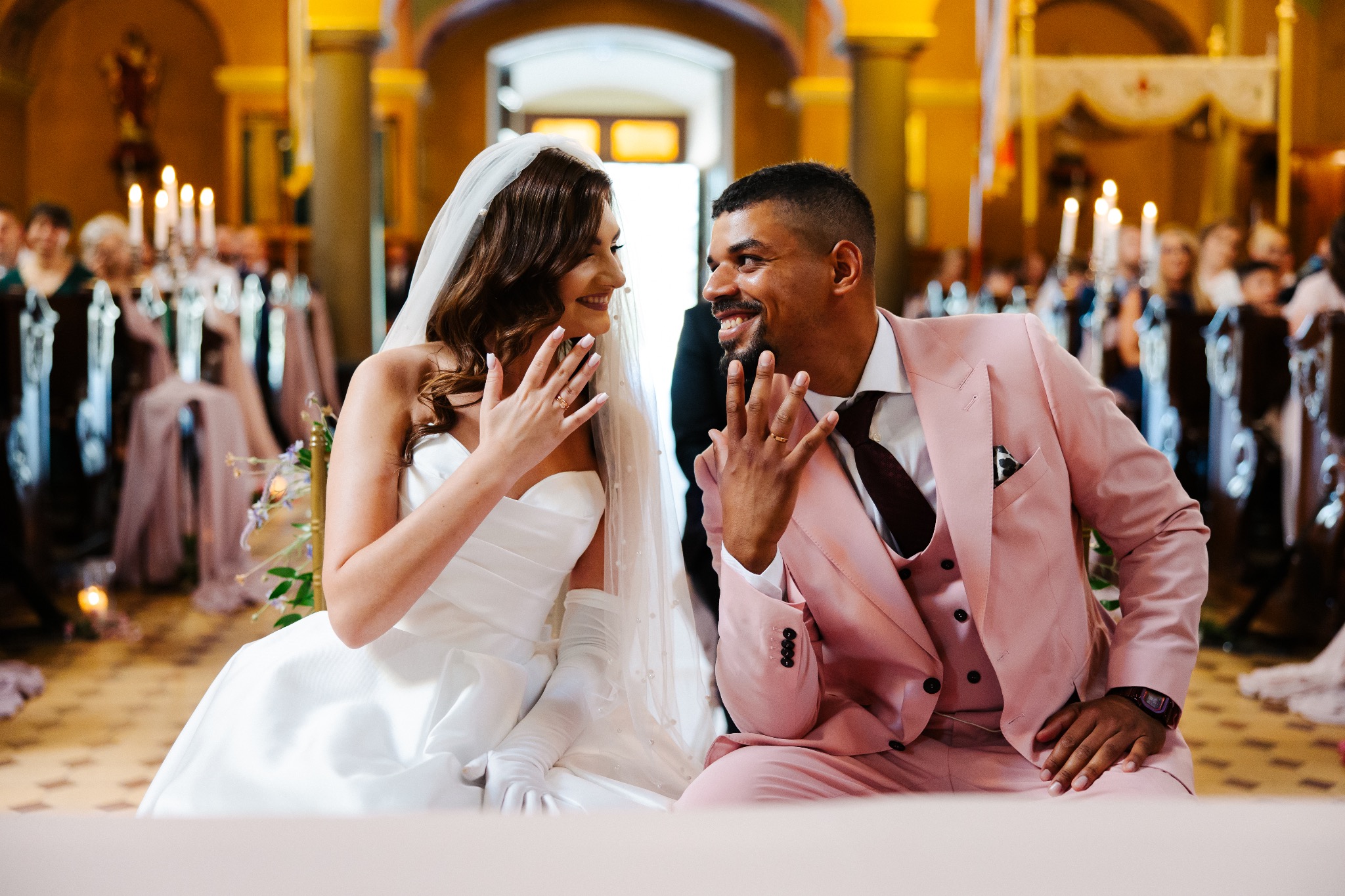 Uśmiechnięta para młoda, w jasnych strojach, siedzi w kościele i prezentuje obrączki. Panna młoda w welonie, pan młody w różowym garniturze. W tle goście i świece.