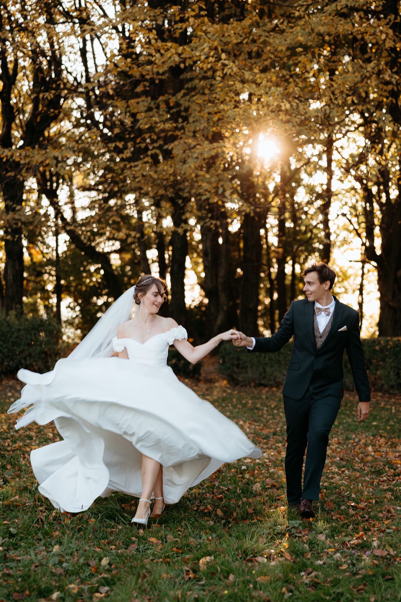 Uśmiechnięta panna młoda w tańcu, wirująca suknia, trzymana za rękę przez pana młodego w zielonym garniturze, jesienne drzewa w tle, ciepłe promienie słońca.