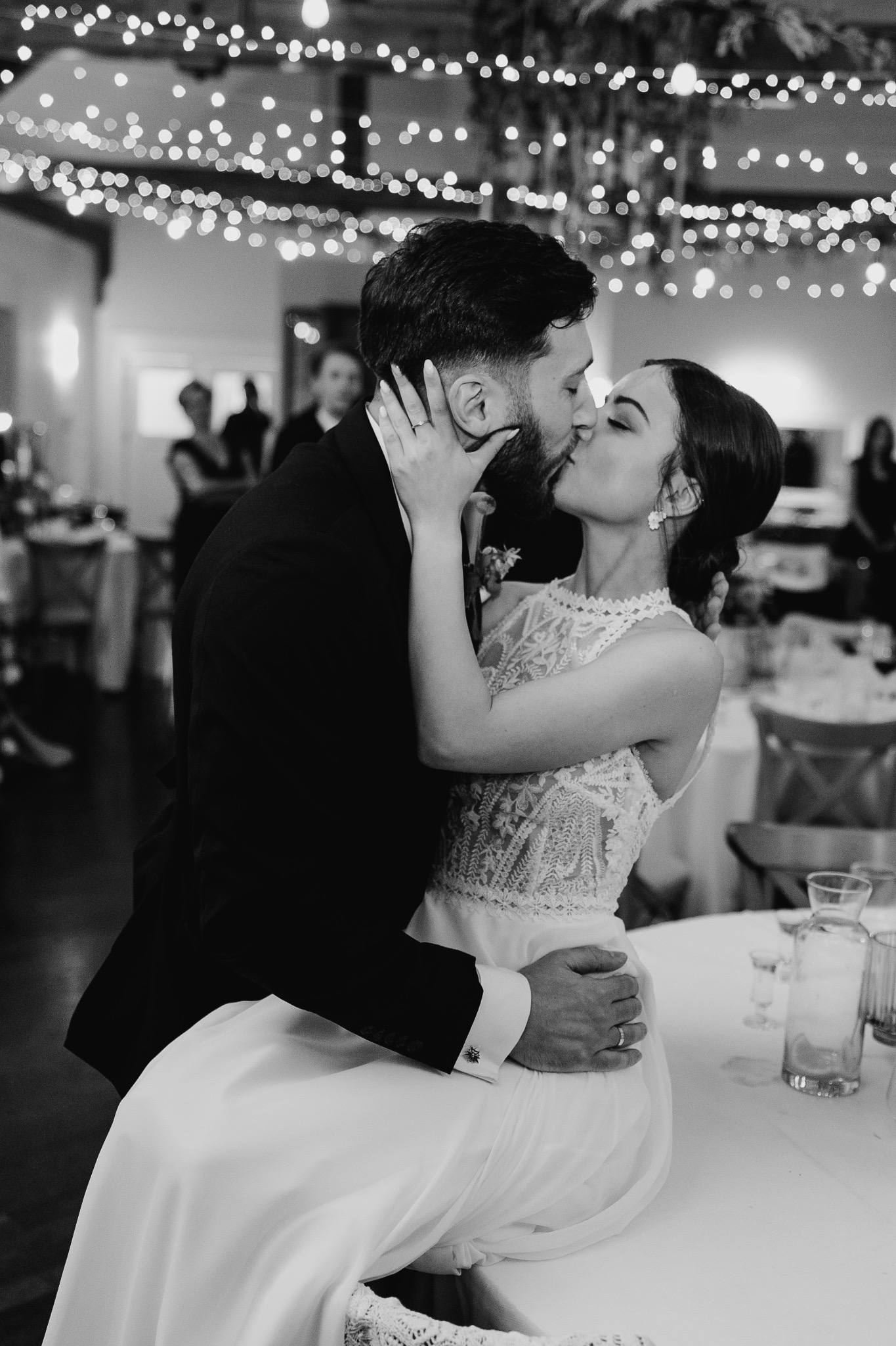 Czarno-białe ujęcie całującej się pary młodej na tle sali weselnej z dekoracyjnym oświetleniem. Panna młoda w koronkowej sukni, pan młody w ciemnym garniturze.