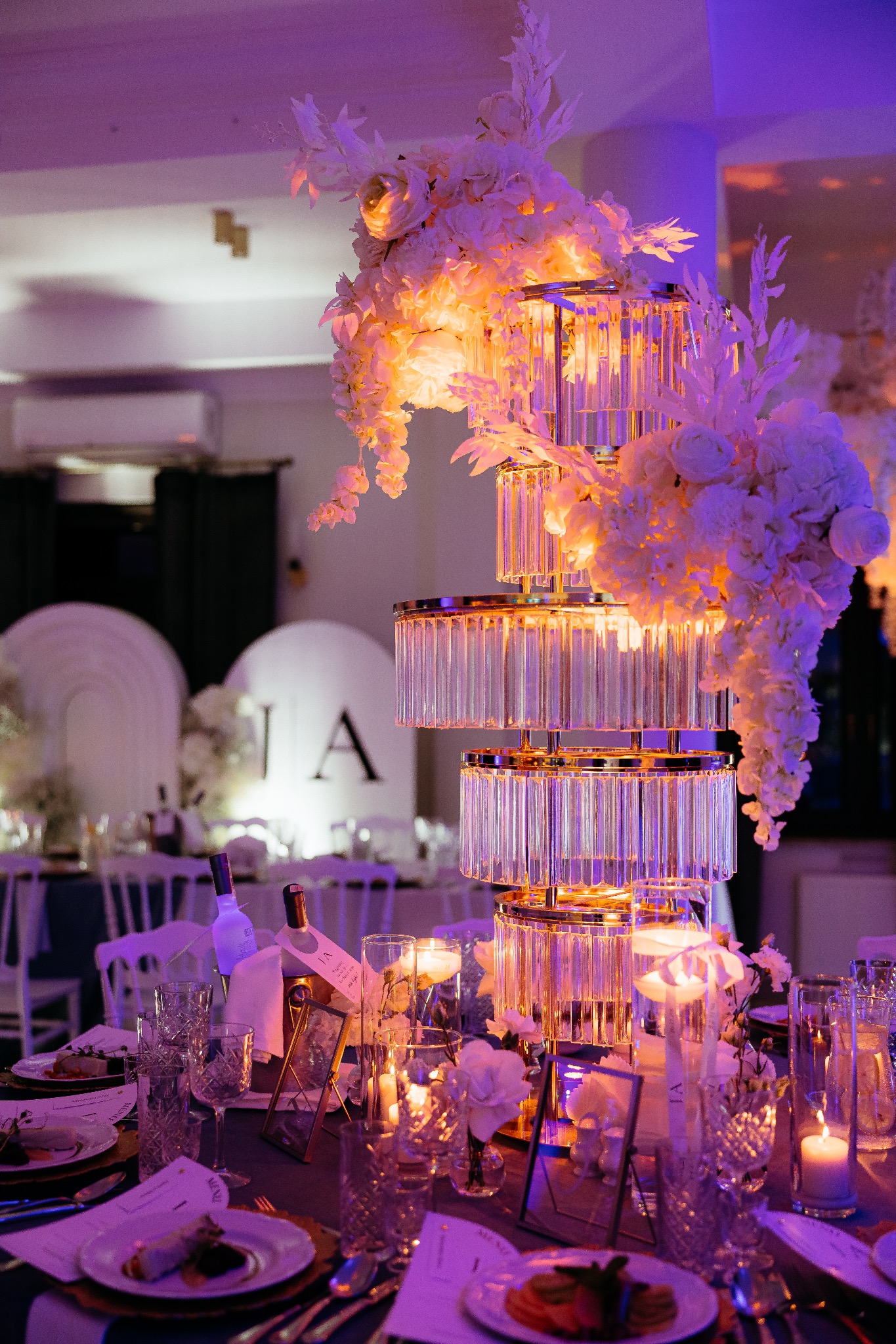 Elegancki stół weselny z kryształową dekoracją kwiatową i świecami, w tle okrągłe elementy dekoracyjne z inicjałami, oświetlony fioletowym światłem.