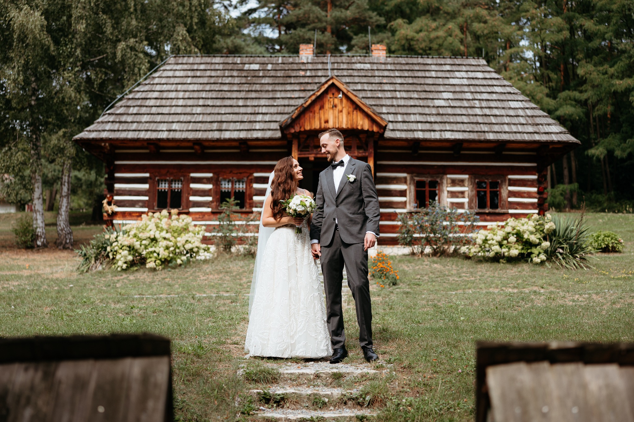 Uśmiechnięta para młoda w plenerze przed drewnianym domem z brązowym dachem i białymi zdobieniami. Panna młoda trzyma bukiet.