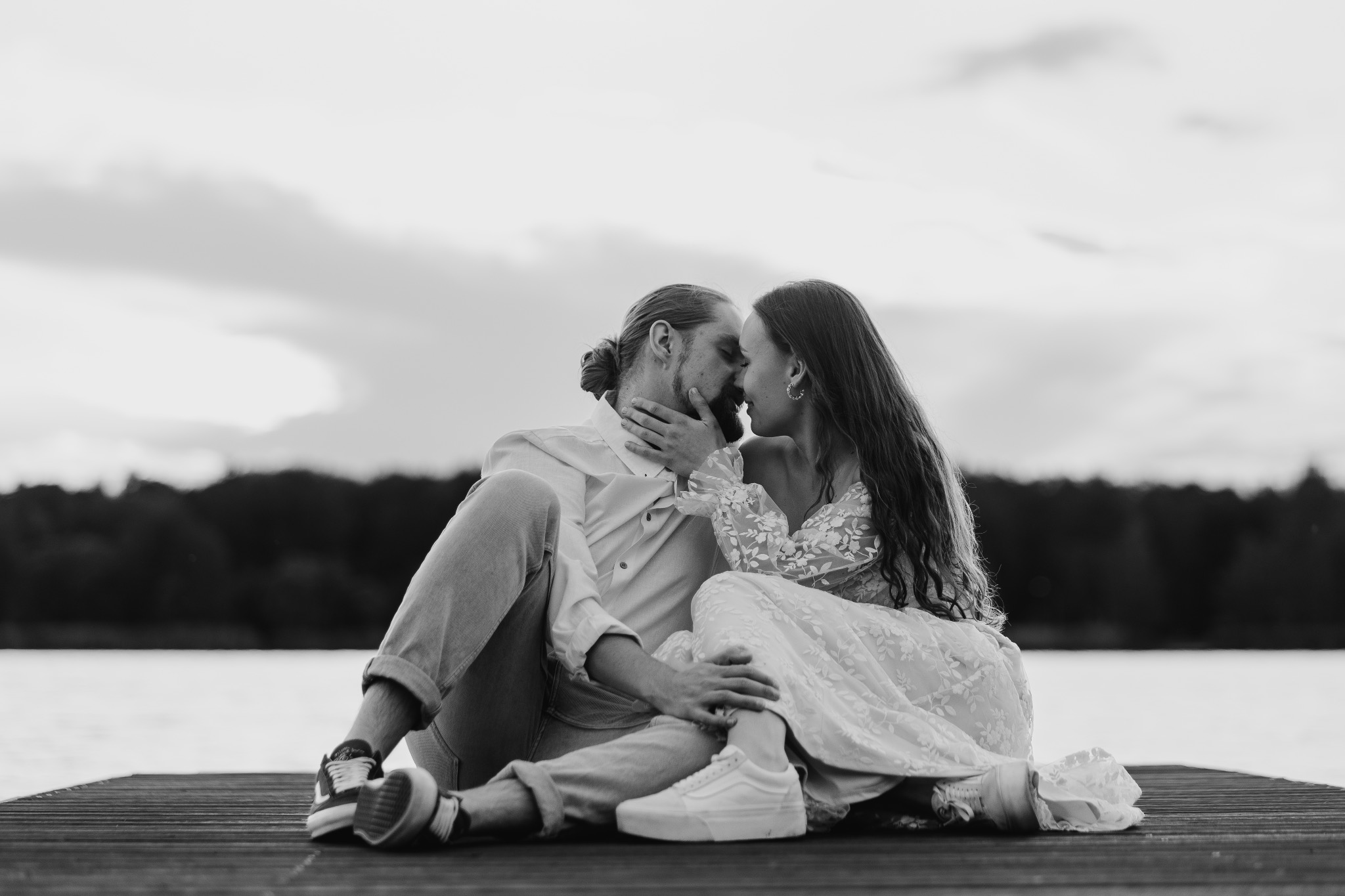 Czarno-białe zdjęcie pary w czułym uścisku na drewnianym pomoście nad jeziorem. Ona w koronkowej sukni, on w koszuli, oboje w sportowych butach, z zalesionym brzegiem w tle.
