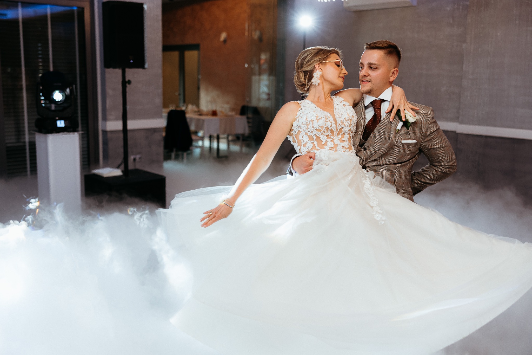 Uśmiechnięta para młoda w tańcu na tle scenicznego dymu i dekoracji sali weselnej. Panna młoda w okularach i sukni z koronką, pan młody w brązowym garniturze w kratę.