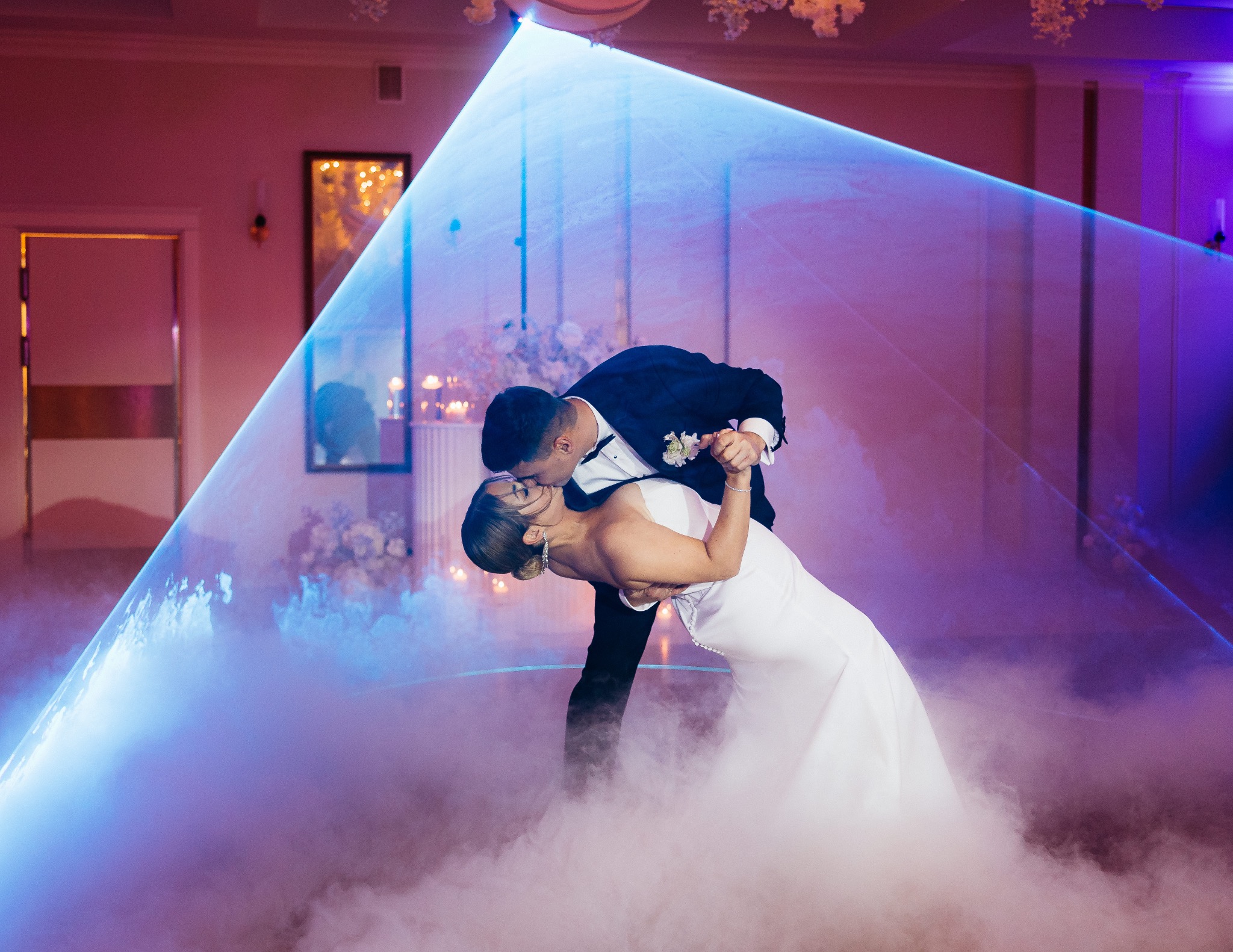 Para młoda w tańcu, otulona mgłą i światłem lasera na sali weselnej. Pan młody w czarnym smokingu, panna młoda w białej sukni. Romantyczna atmosfera.