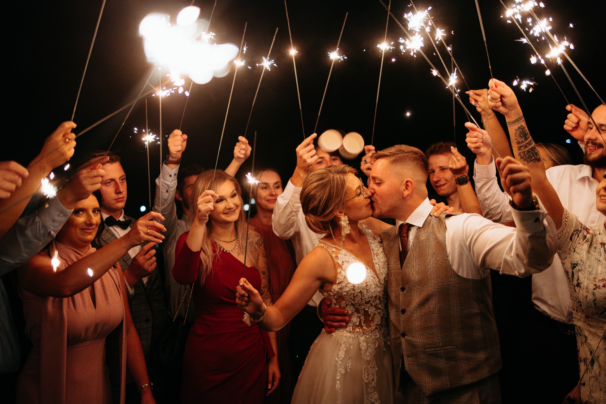 Pocałunek pary młodej otoczonej gośćmi z zimnymi ogniami na tle nocnego nieba. Ujęcie z reportażu ślubnego, z bliska, z efektem bokeh.