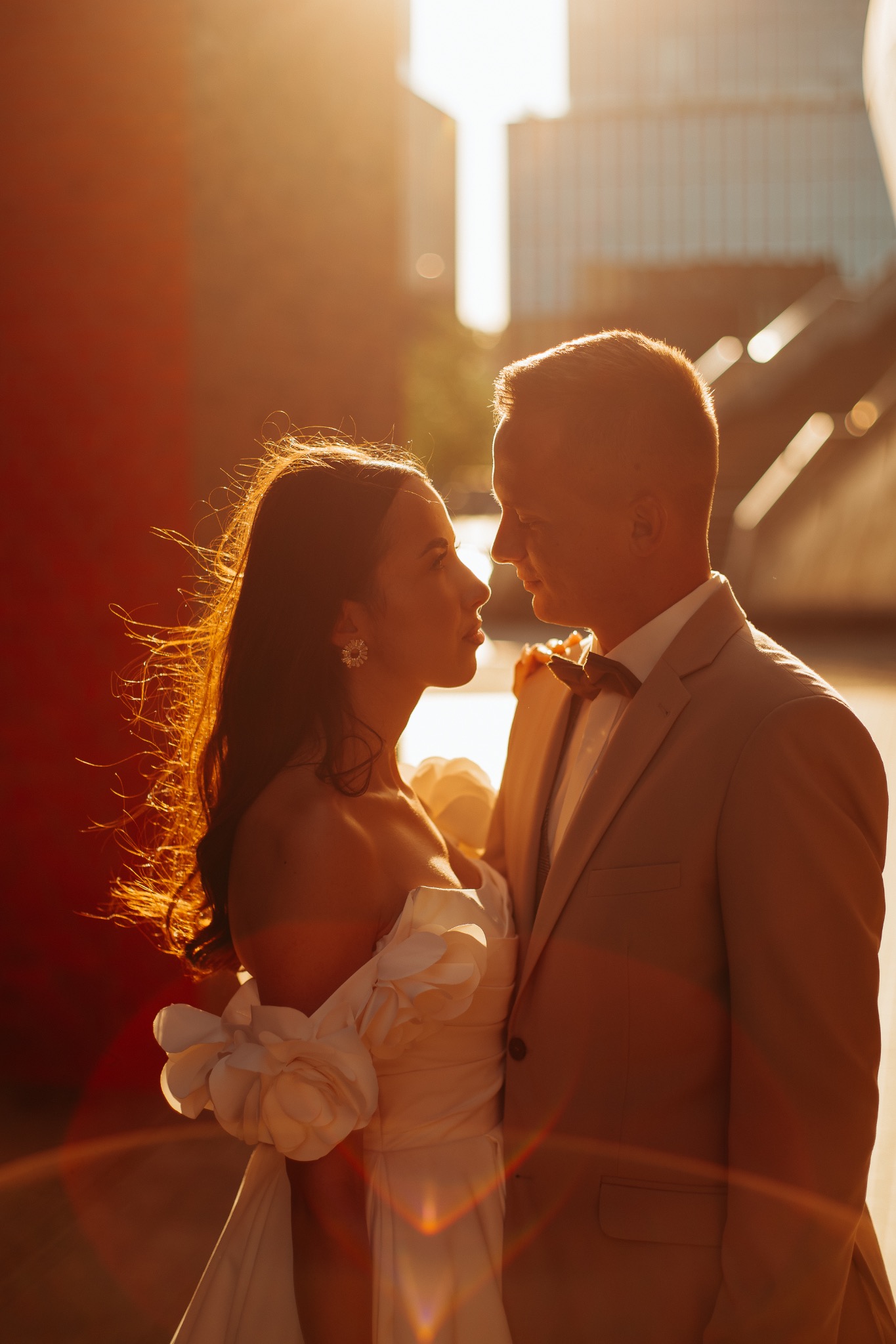 Para młoda w kontraście ciepłego światła: Panna młoda w sukni z kwiatami na ramieniu, pan młody w beżowym garniturze, patrzą na siebie na tle miejskiej architektury.