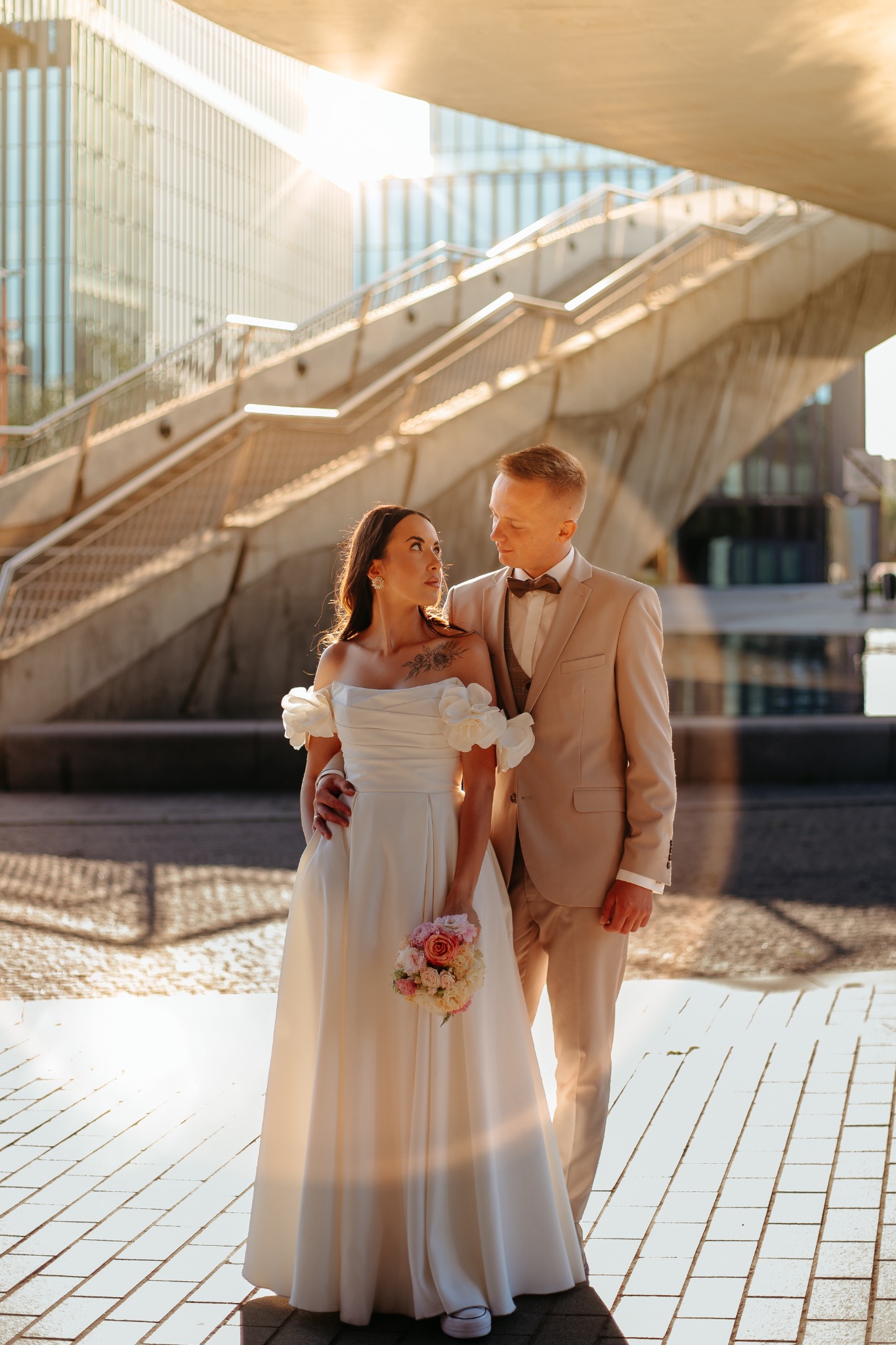 Para młoda w beżowych tonacjach, ona w sukni z kwiatami, on w garniturze, na tle nowoczesnej architektury w słoneczny dzień. Romantyczne spojrzenie, subtelny uśmiech.
