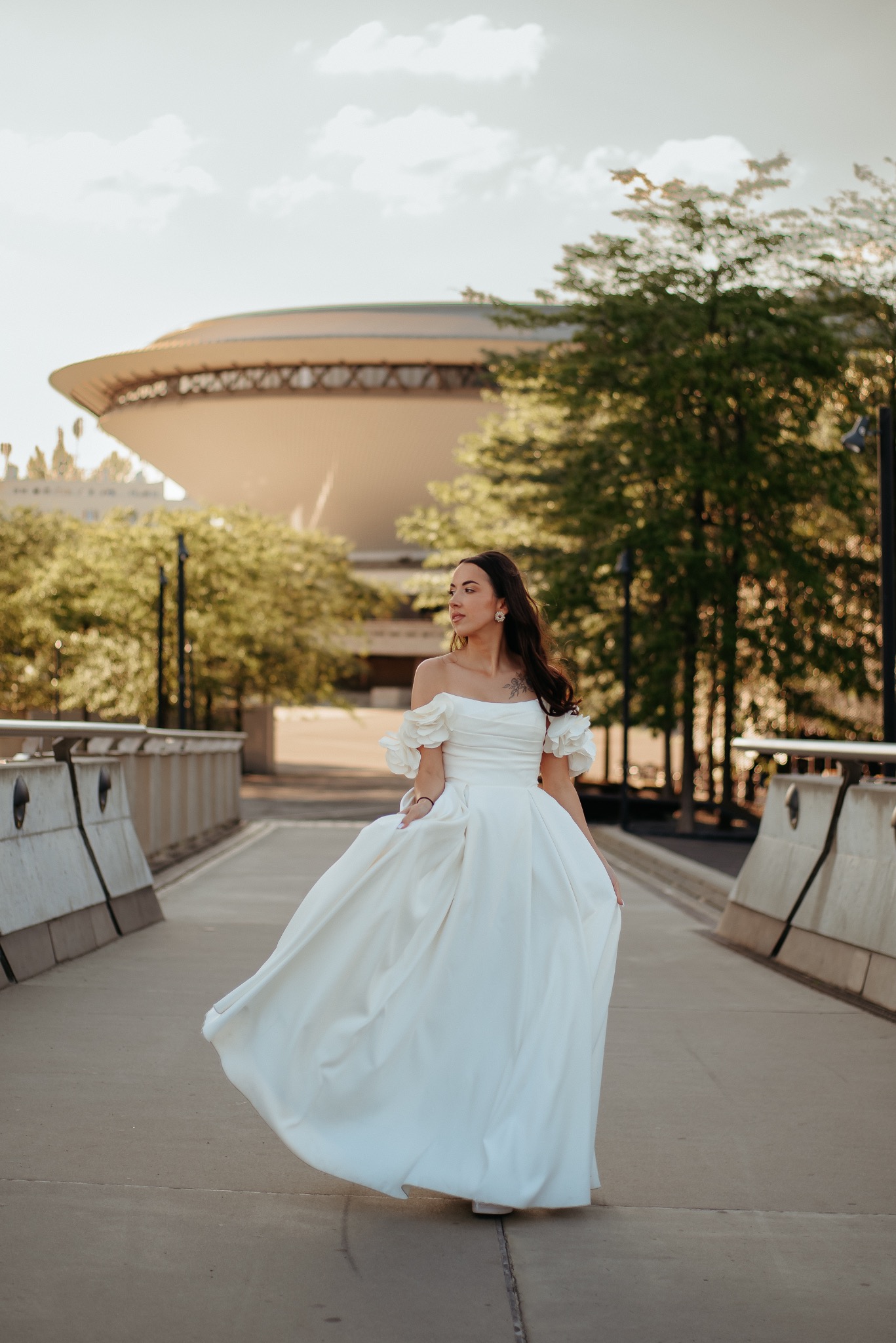 Elegancka sesja ślubna: Panna młoda w białej sukni na moście, w tle Spodek w Katowicach, ciepłe światło zachodzącego słońca, subtelny makijaż i tatuaż na ramieniu.