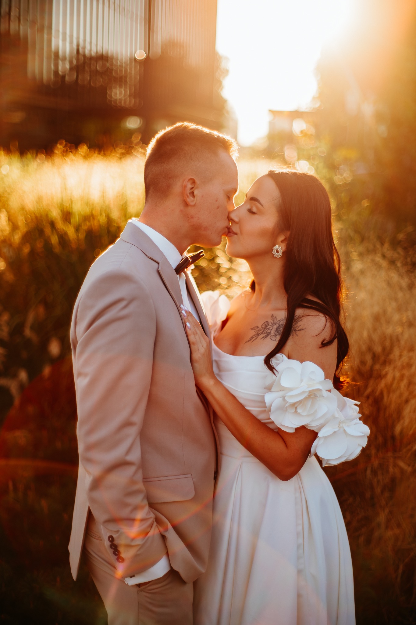 Pocałunek w złocistym świetle: Para młoda w eleganckich strojach, ona w sukni z kwiatowymi aplikacjami, on w beżowym garniturze, na tle wysokich traw.