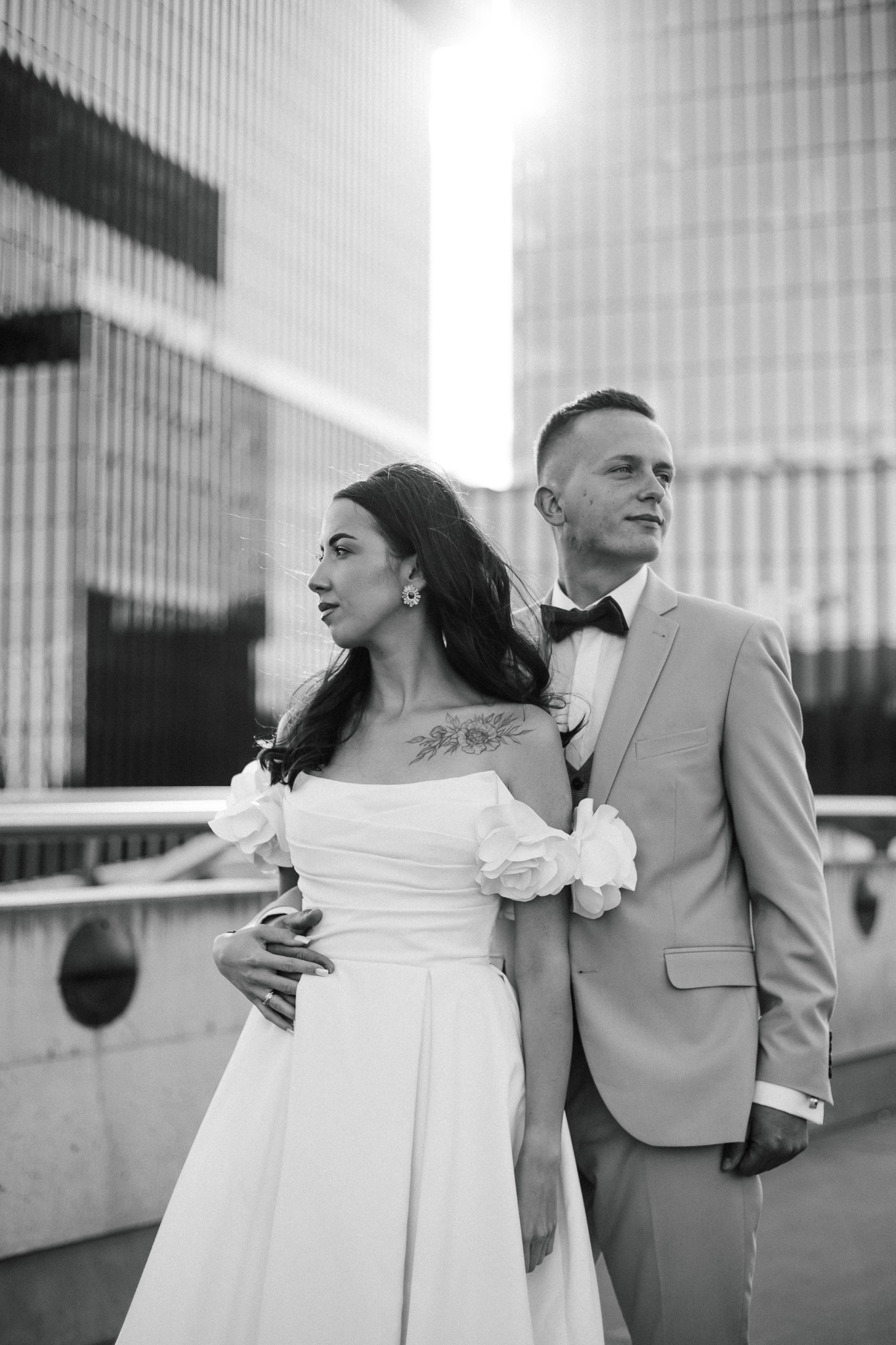 Czarno-białe zdjęcie pary młodej na tle nowoczesnej architektury. Panna młoda w sukni z kwiatami na ramionach, pan młody w garniturze z muchą. Ujęcie w świetle słonecznym.
