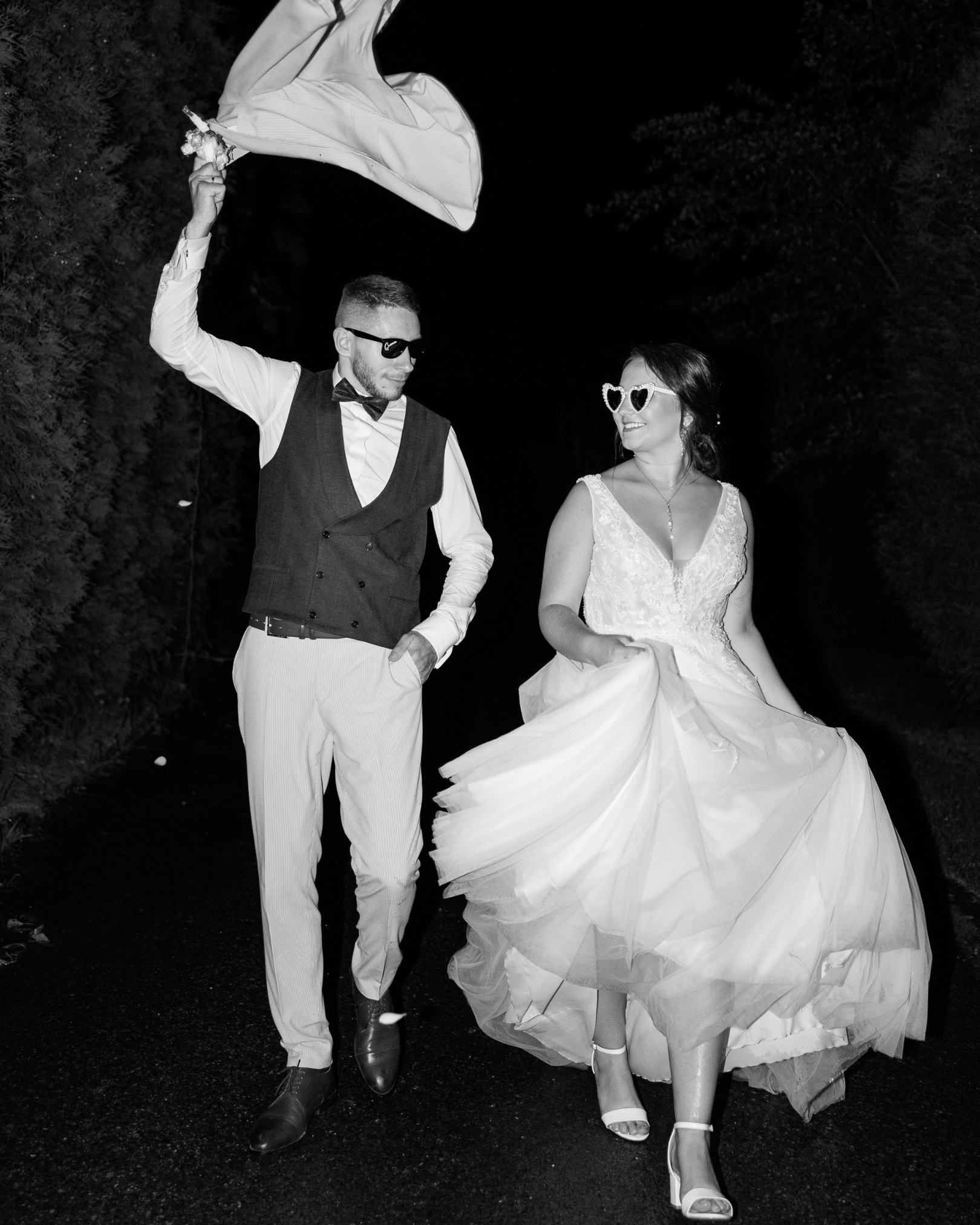 Czarno-białe ujęcie pary młodej w okularach w kształcie serc. Pan młody trzyma marynarkę nad głową. Zdjęcie zrobione w plenerze, nocą.