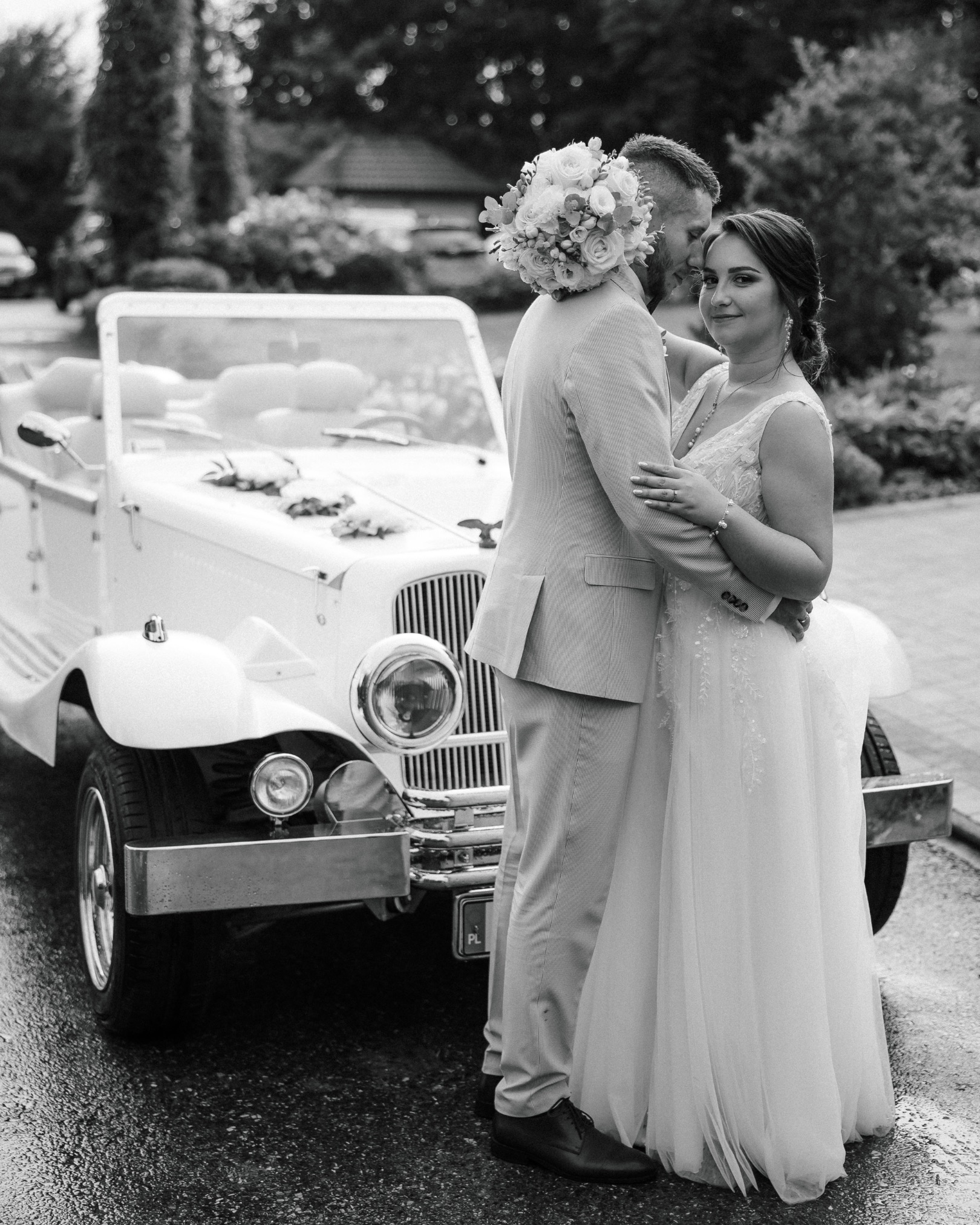 Czarno-białe zdjęcie pary młodej przy zabytkowym kabriolecie, bukiet ślubny na ramieniu pana młodego, uśmiechnięta panna młoda patrzy w obiektyw.